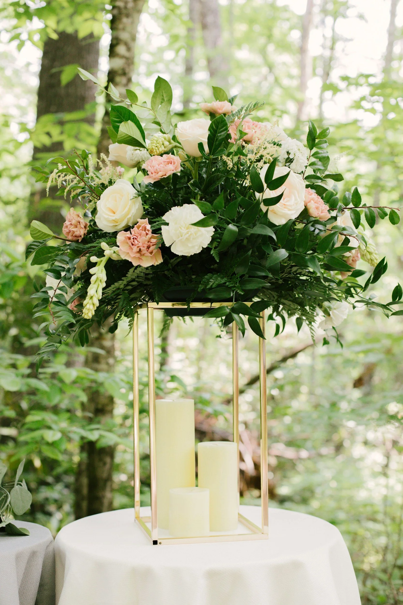 Elegant floral centerpiece with white and light pink flowers on a gold metal frame, holding three white candles, placed on a round table with a white tablecloth, outdoors in a forest setting.