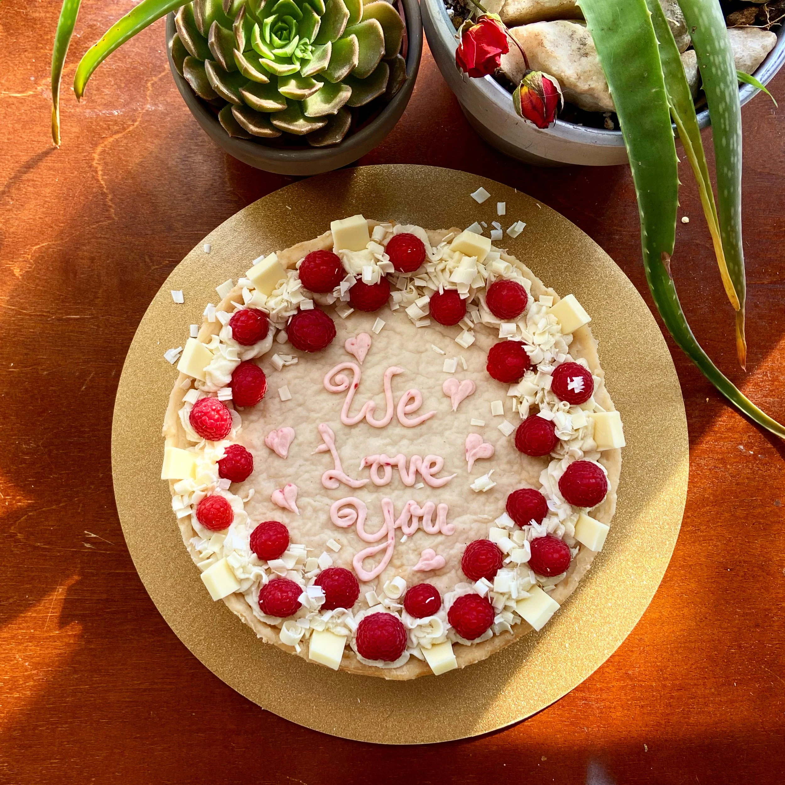 Round cake with white frosting, decorated with raspberries, white chocolate shavings, and white chocolate chunks. Pink lettering on the cake reads "We Love You" surrounded by small pink hearts. The cake is on a gold-colored board on a wooden table, with potted succulents nearby.
