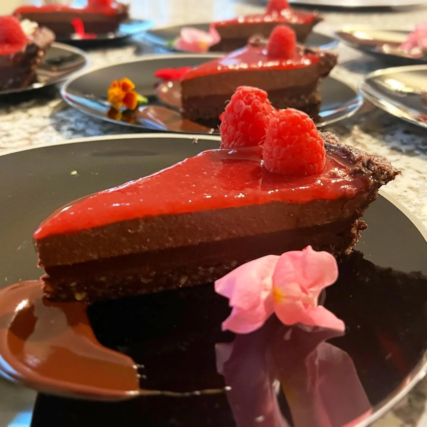 Chocolate dessert with red fruit topping, garnished with raspberries and pink edible flowers on a black plate.