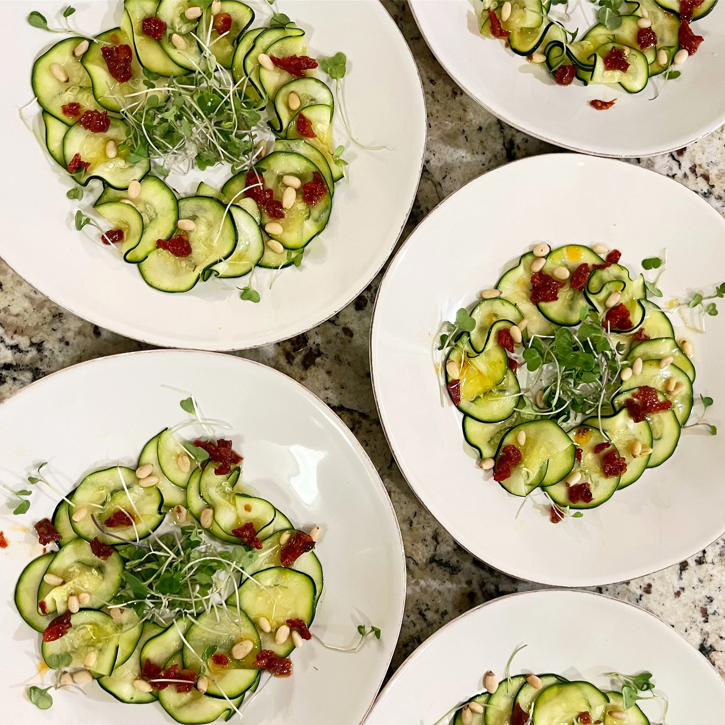 Salad plates with sliced cucumbers, microgreens, pine nuts, and sun-dried tomatoes arranged in a circular pattern on white plates.