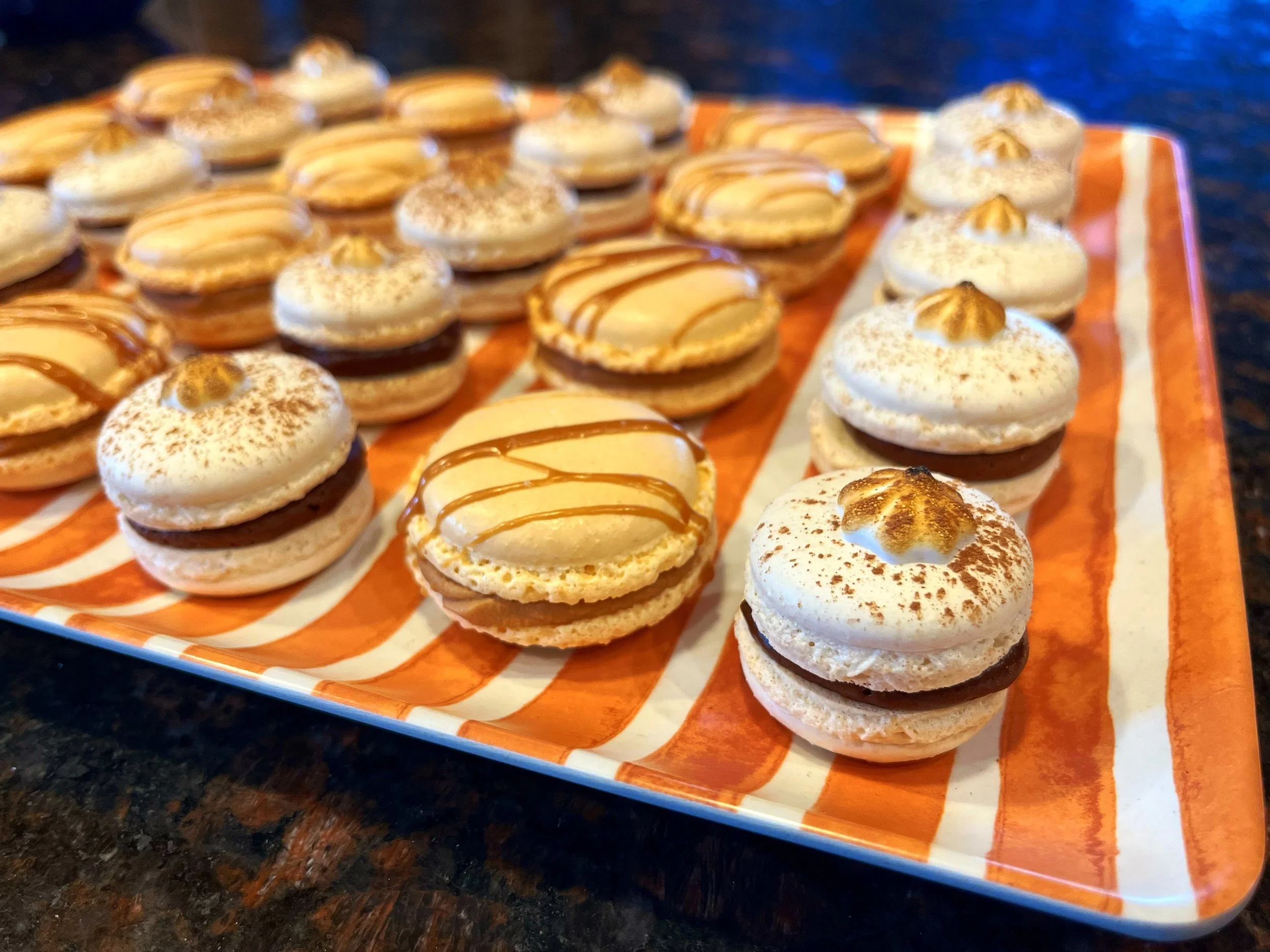 Plate of assorted macarons with white and brown filling, decorated with cocoa powder and caramel drizzle on an orange and white striped dish.