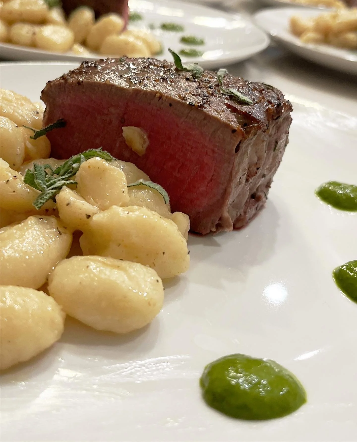 Seared steak with gnocchi pasta garnished with herbs and a drizzle of green sauce on a white plate.