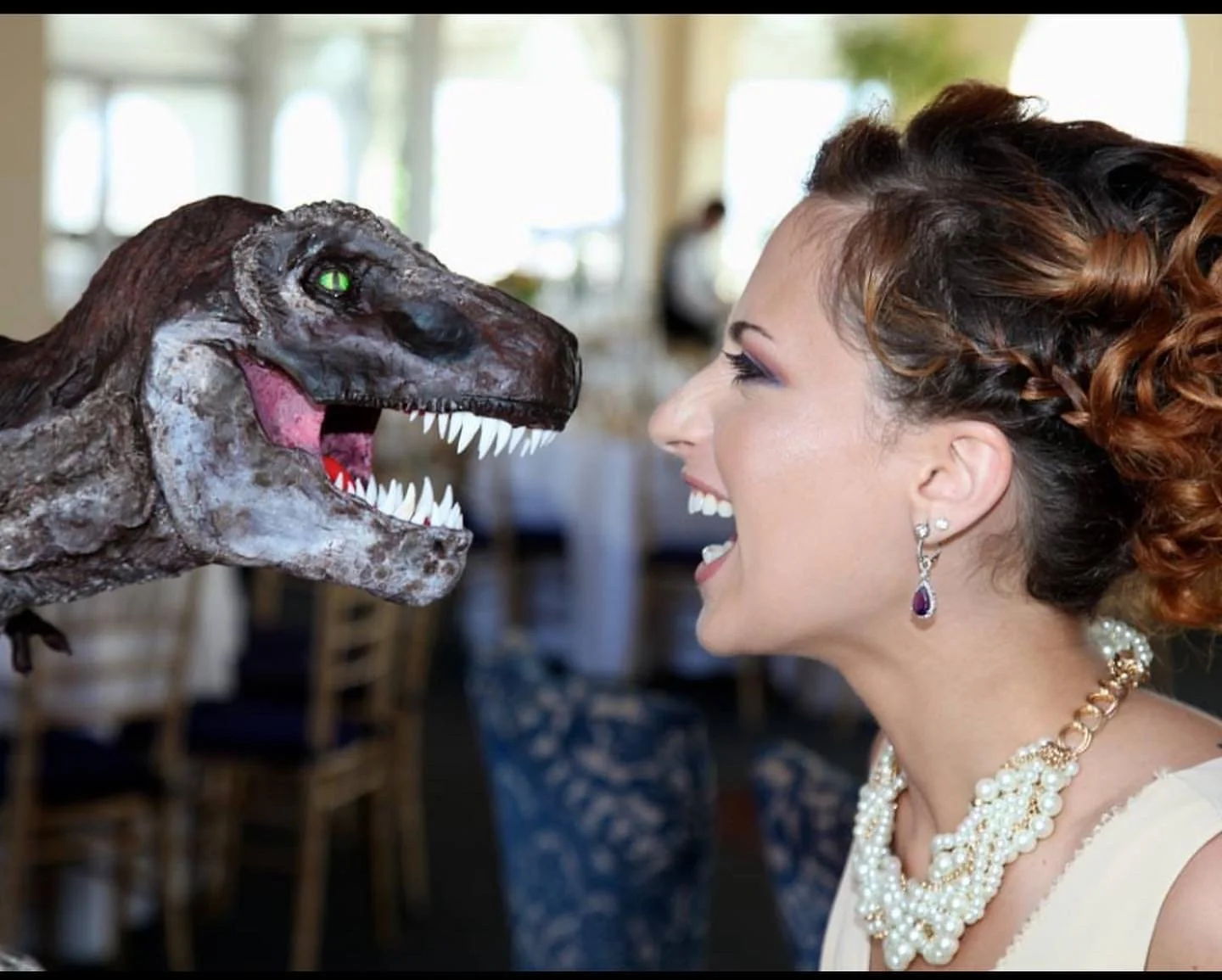 A woman with styled hair, pearl necklace, and earrings, smiling as she faces a life-sized dinosaur model with sharp teeth and green eyes.