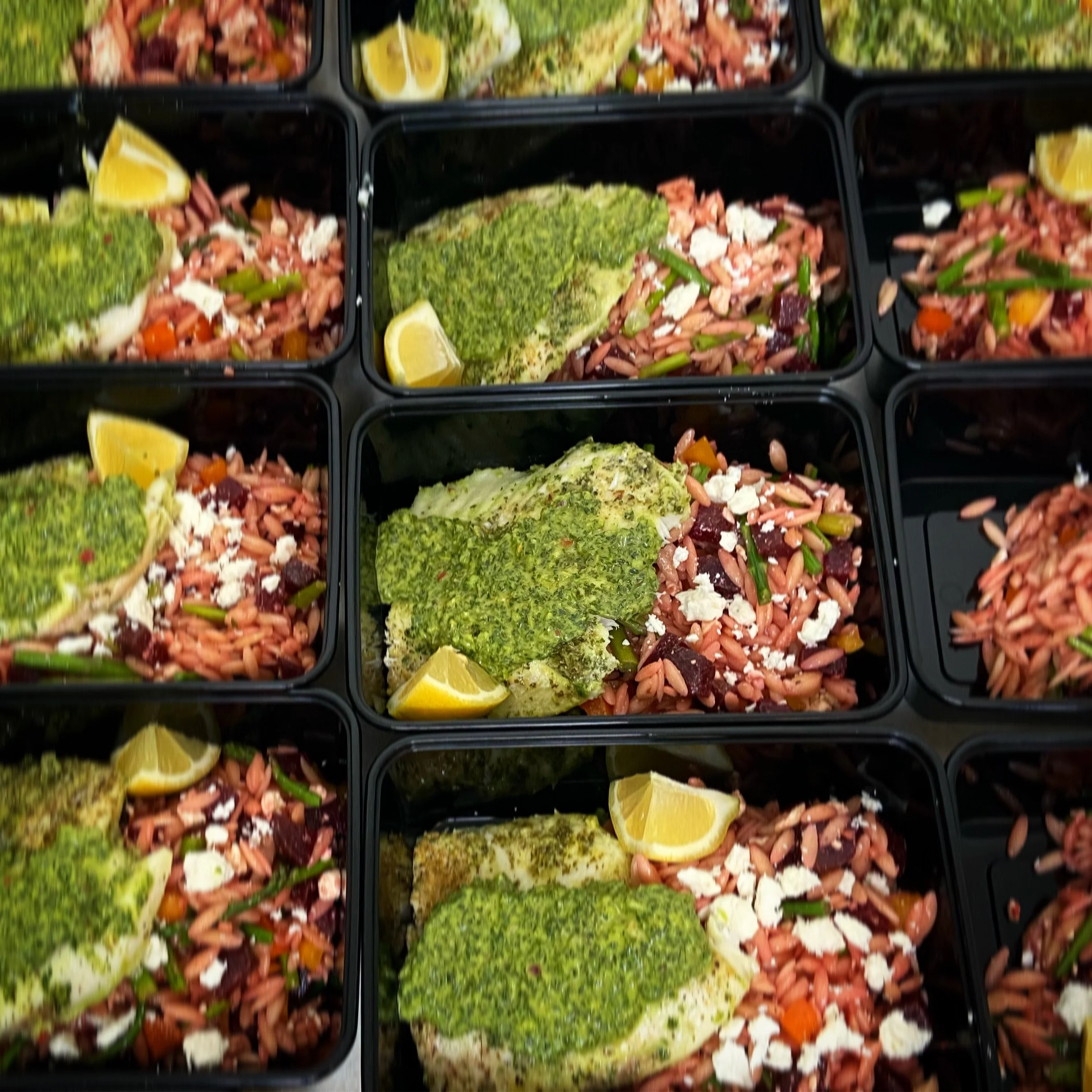 Multiple black meal prep containers with baked fish topped with green pesto, lemon wedges, and a side of rice with vegetables.