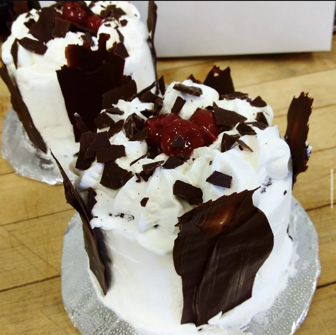 Two slices of cake topped with whipped cream, chocolate shavings, and cherries on a wooden surface.