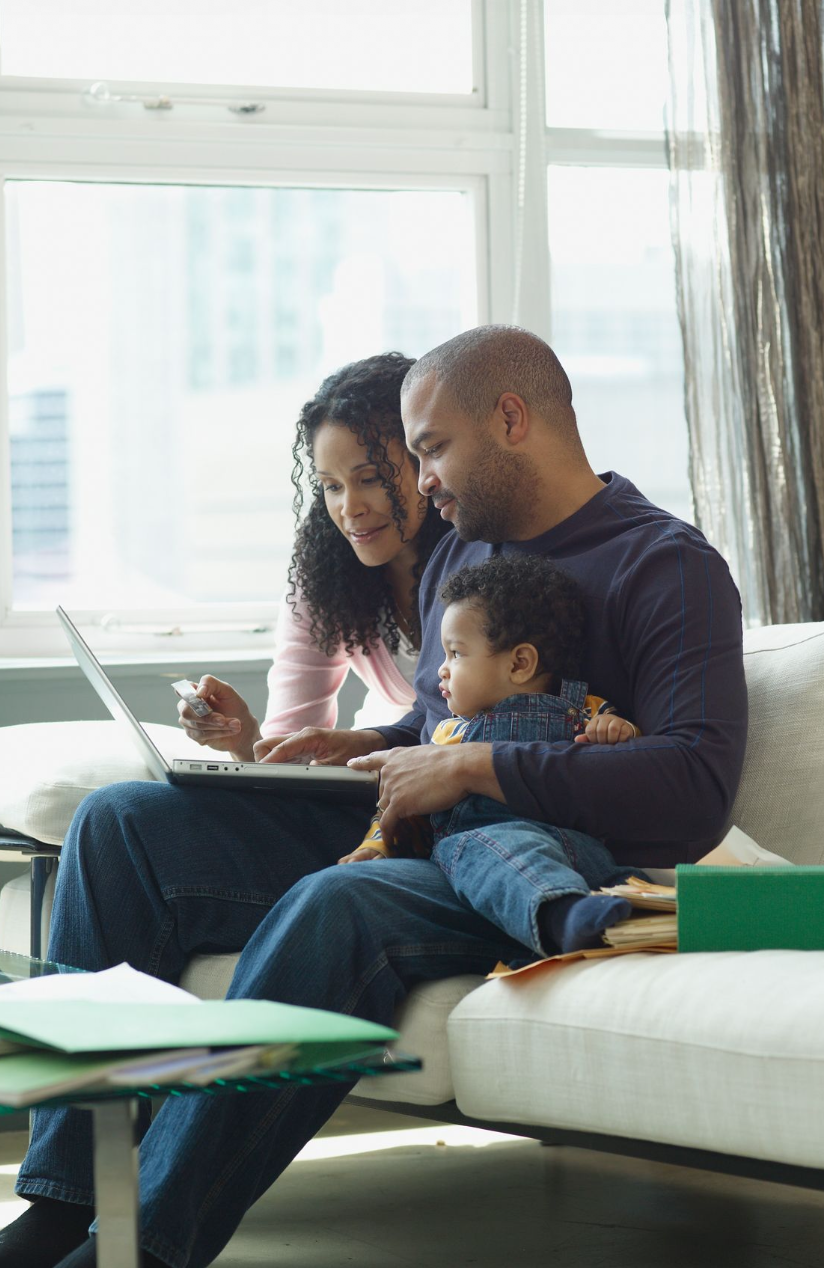 A family of three sitting on a couch in a bright living room, looking at a laptop together. The father is holding a young child, and the mother is leaning in, pointing at the laptop screen.