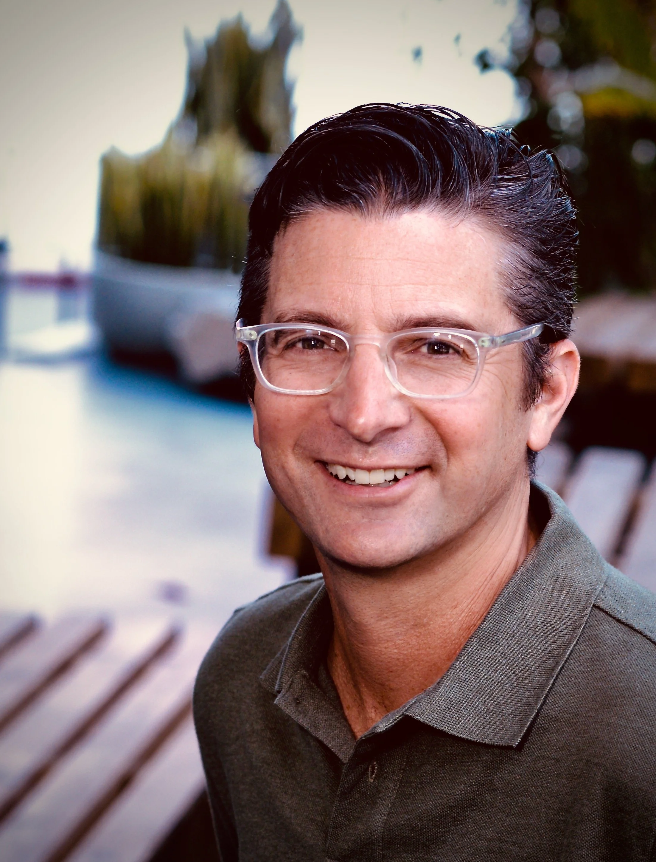 A man with dark hair, wearing glasses, and a brown polo shirt, smiling outdoors near a wooden bench, with decorative plants in the background.