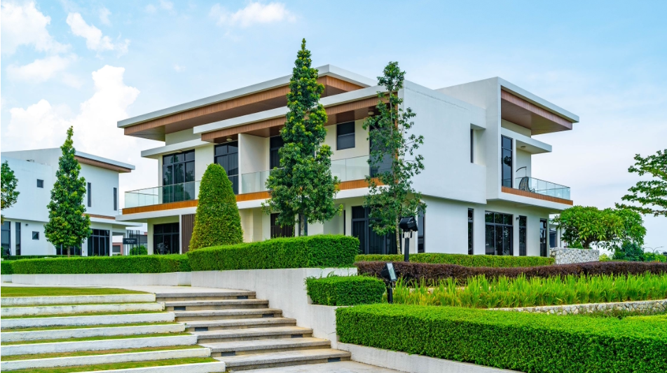 Modern two-story white house with flat roof, large windows, and balconies, surrounded by well-manicured greenery and trees, with a concrete staircase leading up to the entrance.