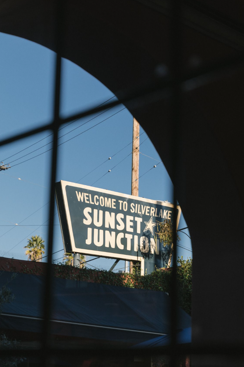 A road sign reading 'Welcome to Silverlake Sunset Junction' with a star symbol, viewed through a window with a grid pattern, against a clear blue sky with utility poles and palm trees in the background.
