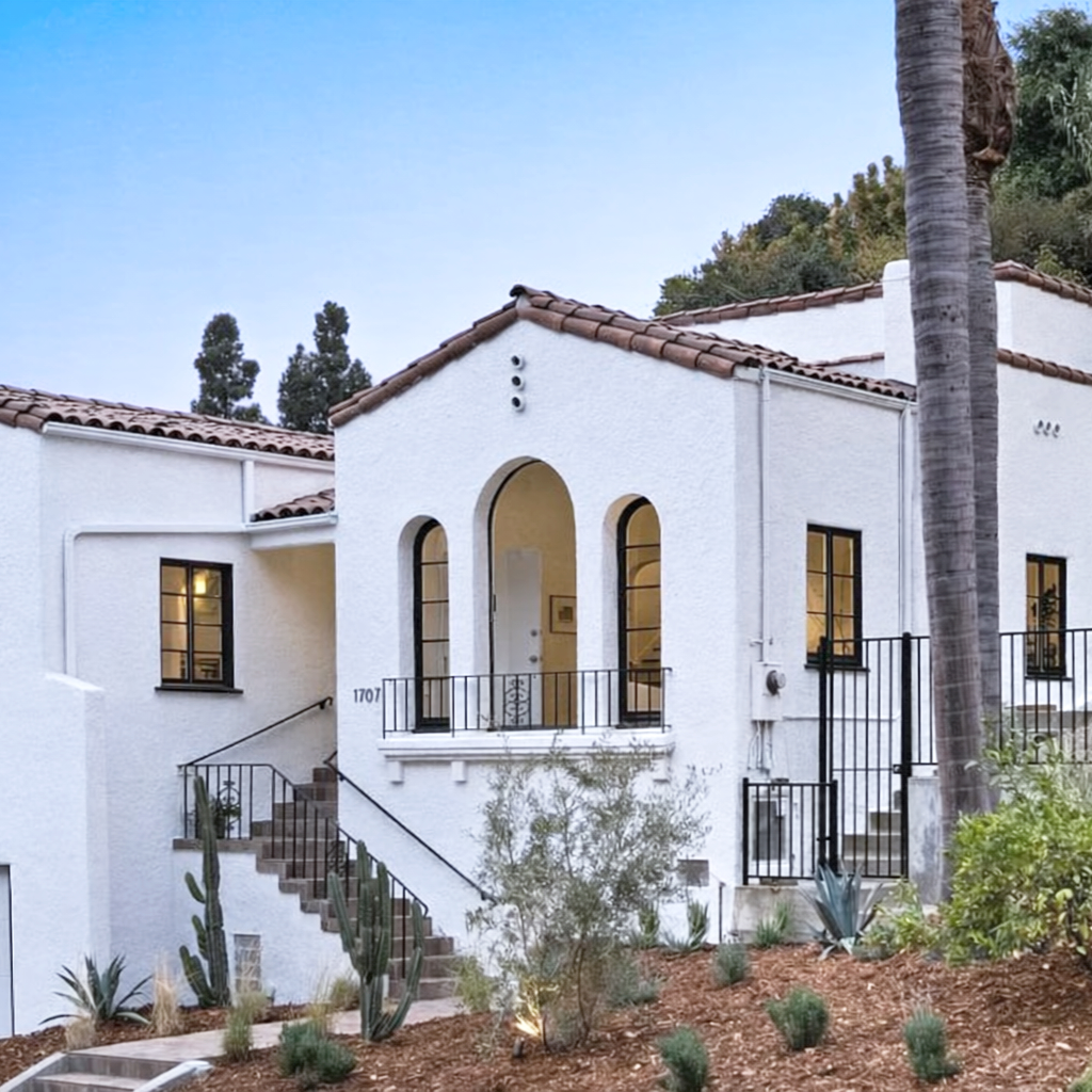 A white stucco house with Spanish-style red tile roof, arched doorway, and black metal railings, surrounded by desert plants and tall palm trees, during twilight.