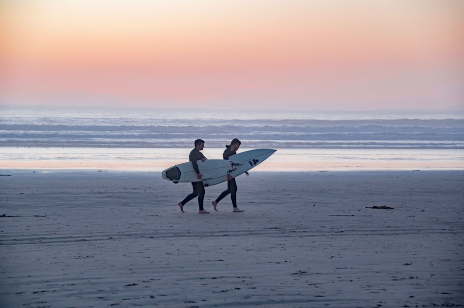 Two people walking on the beach at sunset, carrying a surfboard.