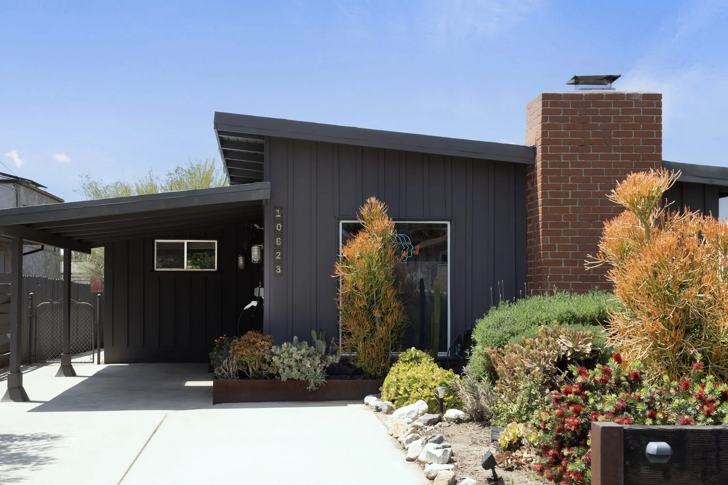 A modern house with black exterior, a brick chimney, and a landscaped front yard with various plants and succulents.