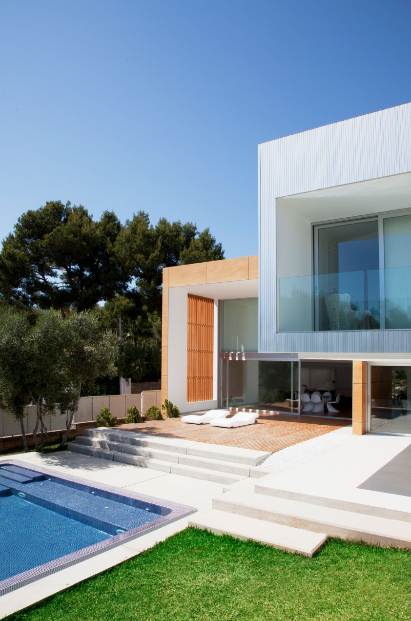 Modern house with a swimming pool in the backyard, white steps leading to the patio, outdoor lounge chairs, glass sliding doors, and trees in the background.