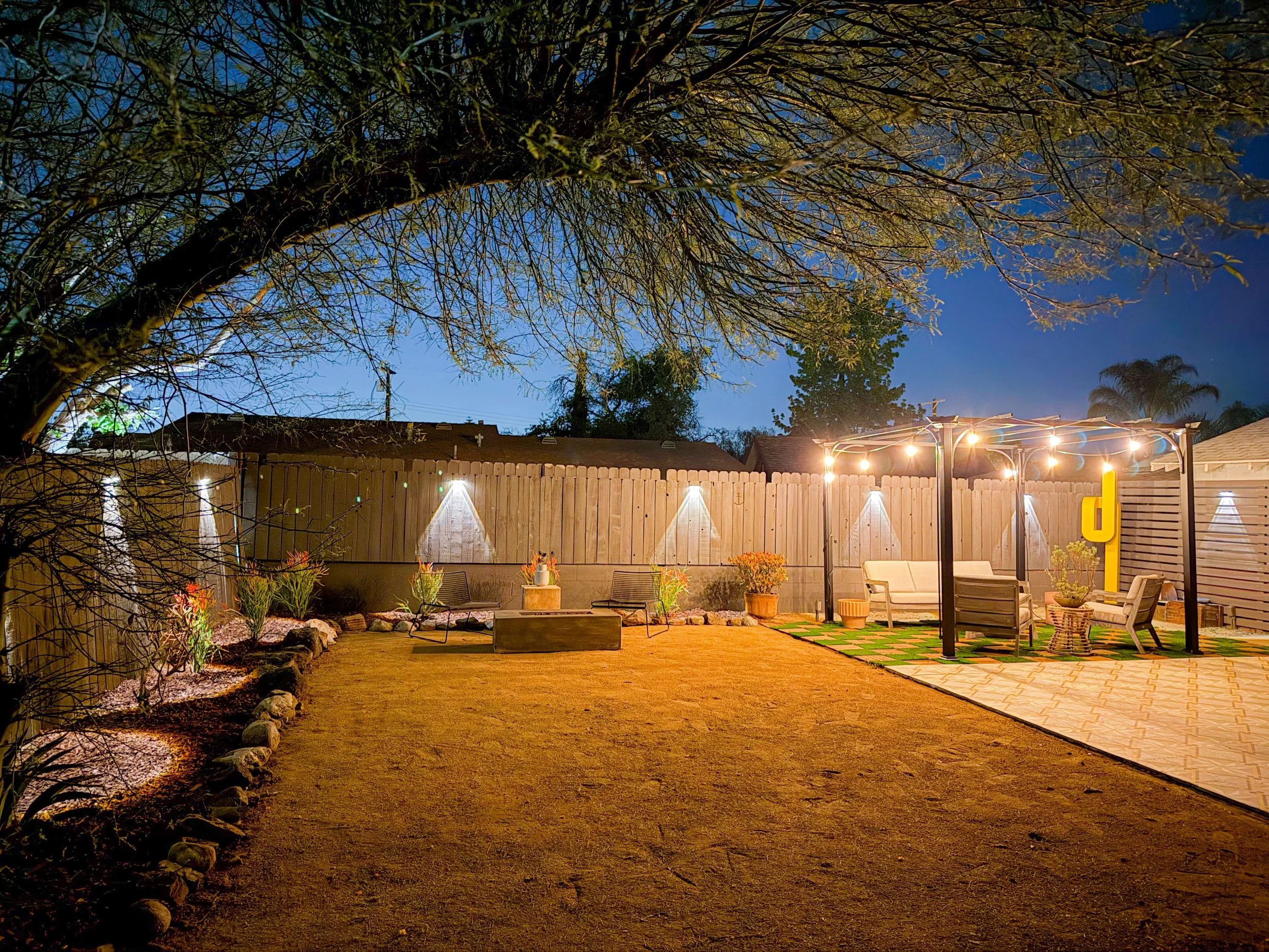 A backyard patio at night with string lights hanging over a seating area, surrounded by a wooden privacy fence, trees, and potted plants.