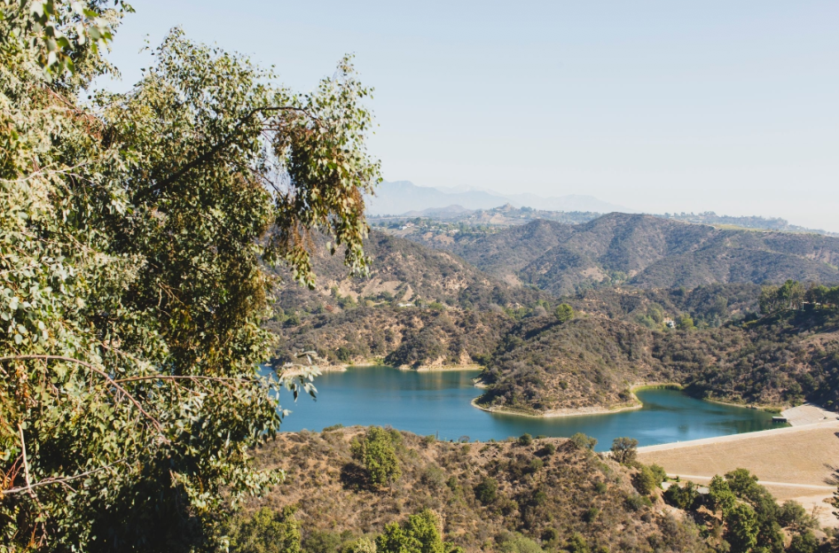 A landscape view of a reservoir surrounded by hills and mountains with sparse vegetation, under a clear sky.