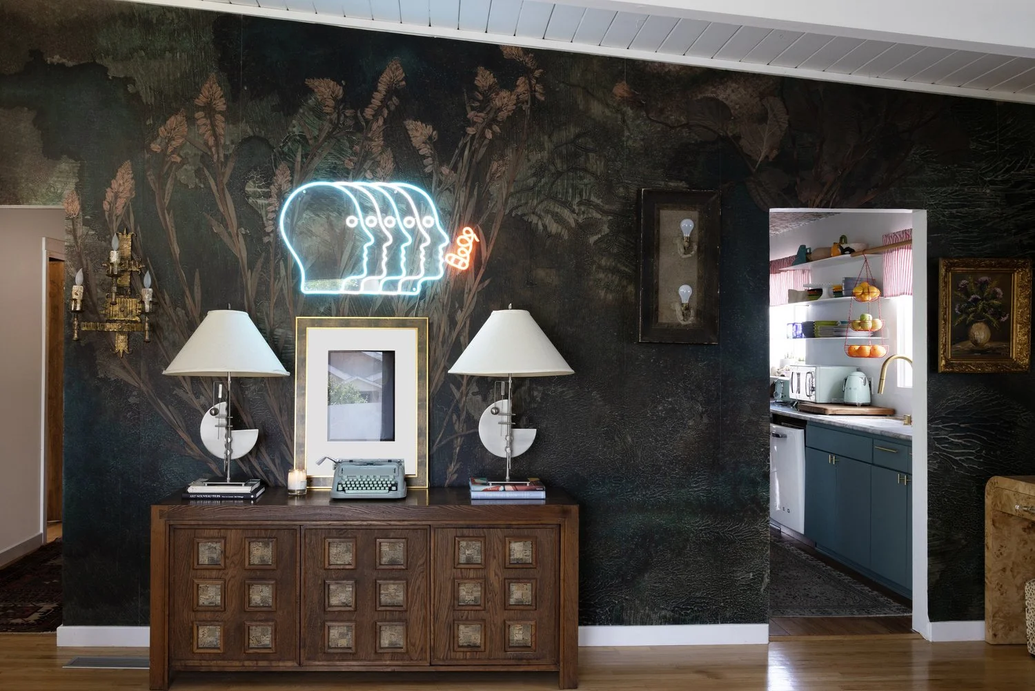 A living room wall with a dark botanical wallpaper, neon signs of four overlapping human profiles and a butterfly, a gold-framed mirror, two table lamps, a vintage typewriter, and framed art. To the right, an opening reveals a kitchen with teal cabinets, open shelves with dishes, a toaster, kettle, and a window with pink striped curtains.