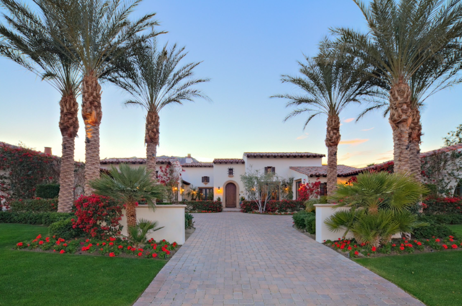 A luxurious villa with a stone pathway leading to the entrance, surrounded by palm trees, colorful flowers, and well-maintained lawn under a clear sky at sunset.