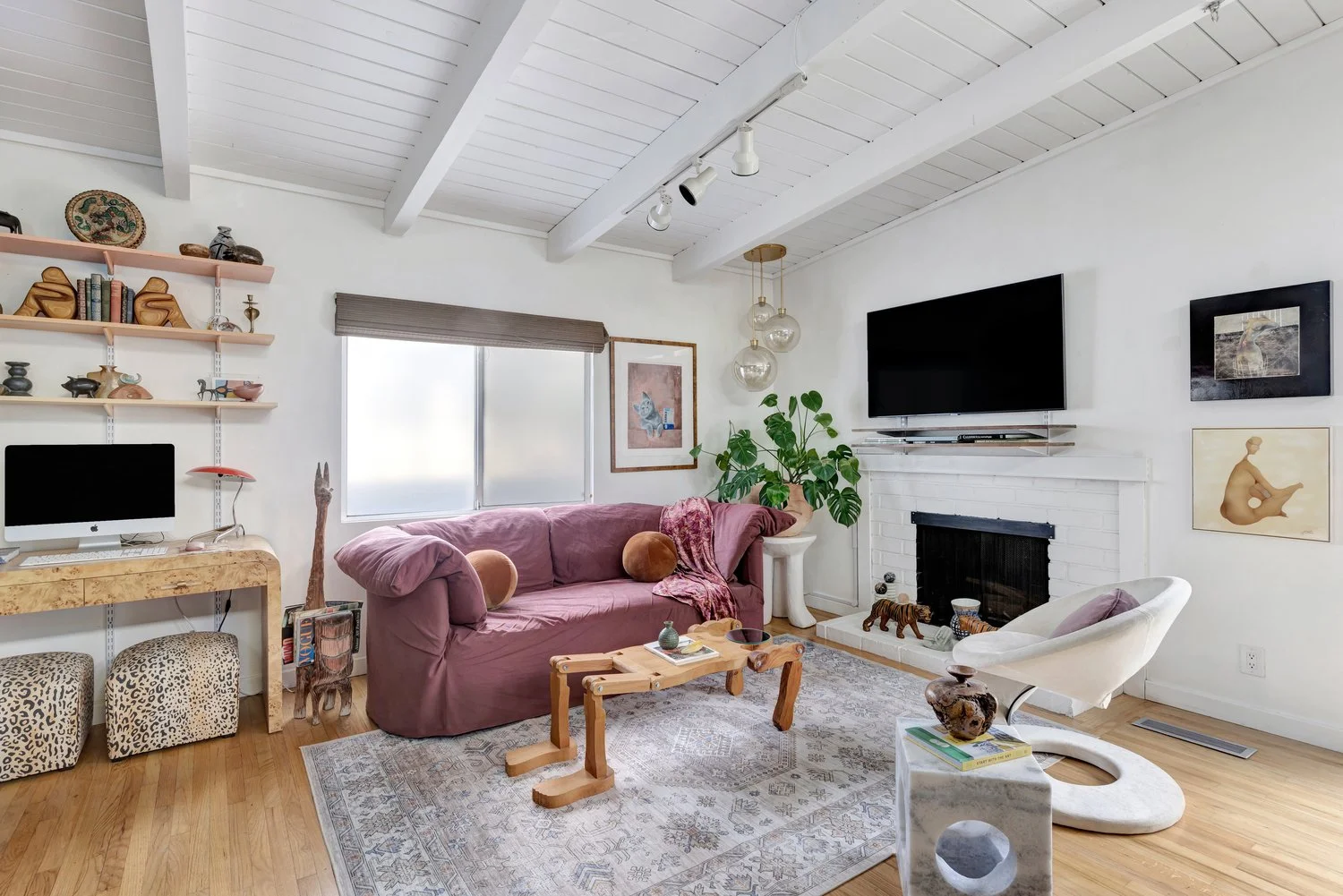 Living room with vaulted white ceiling, large window, pink sofa with pillows, modern chair, fireplace, TV, open shelves with decor, potted plant, artwork, and area rug.