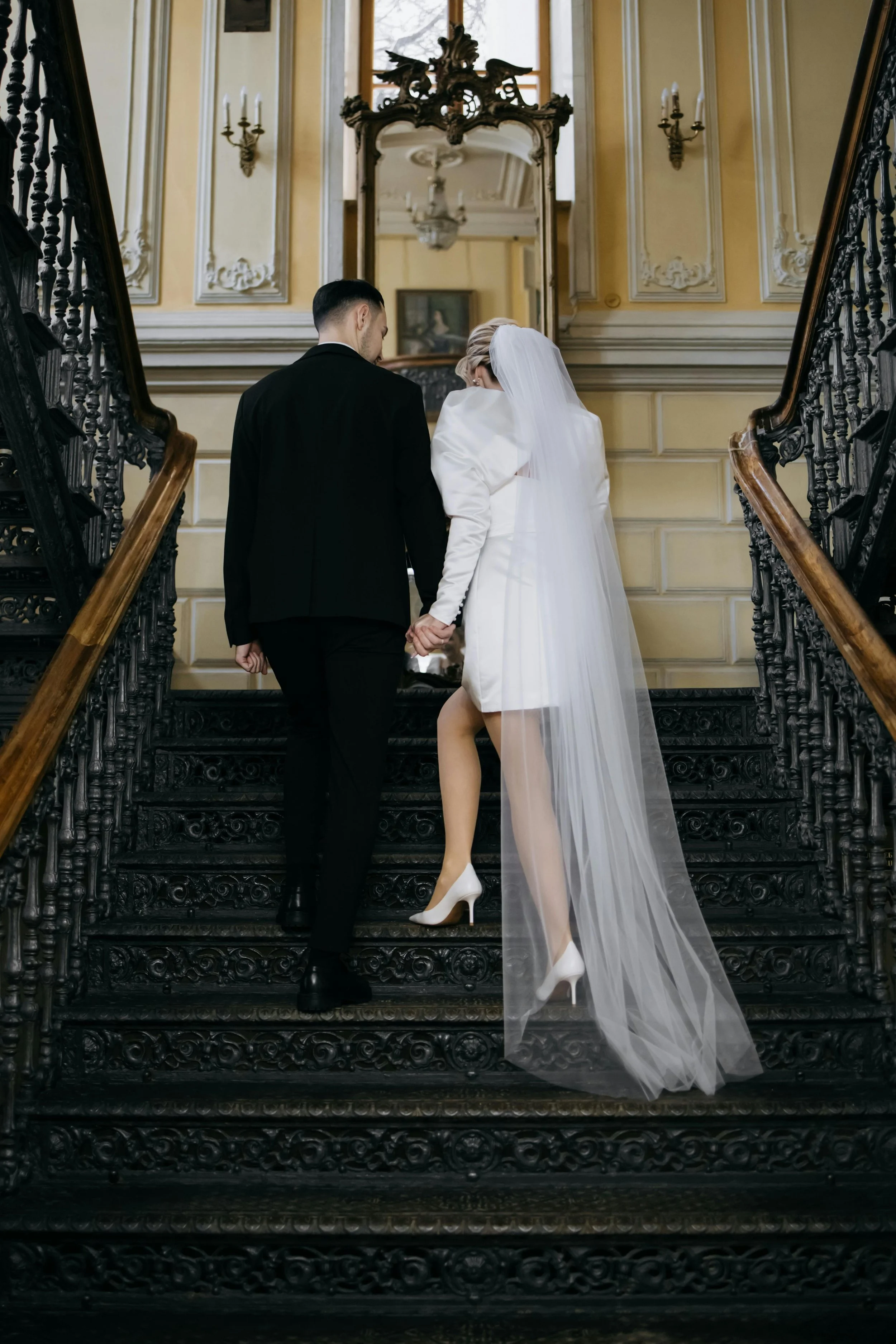 A bride and groom holding hands as they ascend a dark, ornate staircase in a grand, vintage-style building.