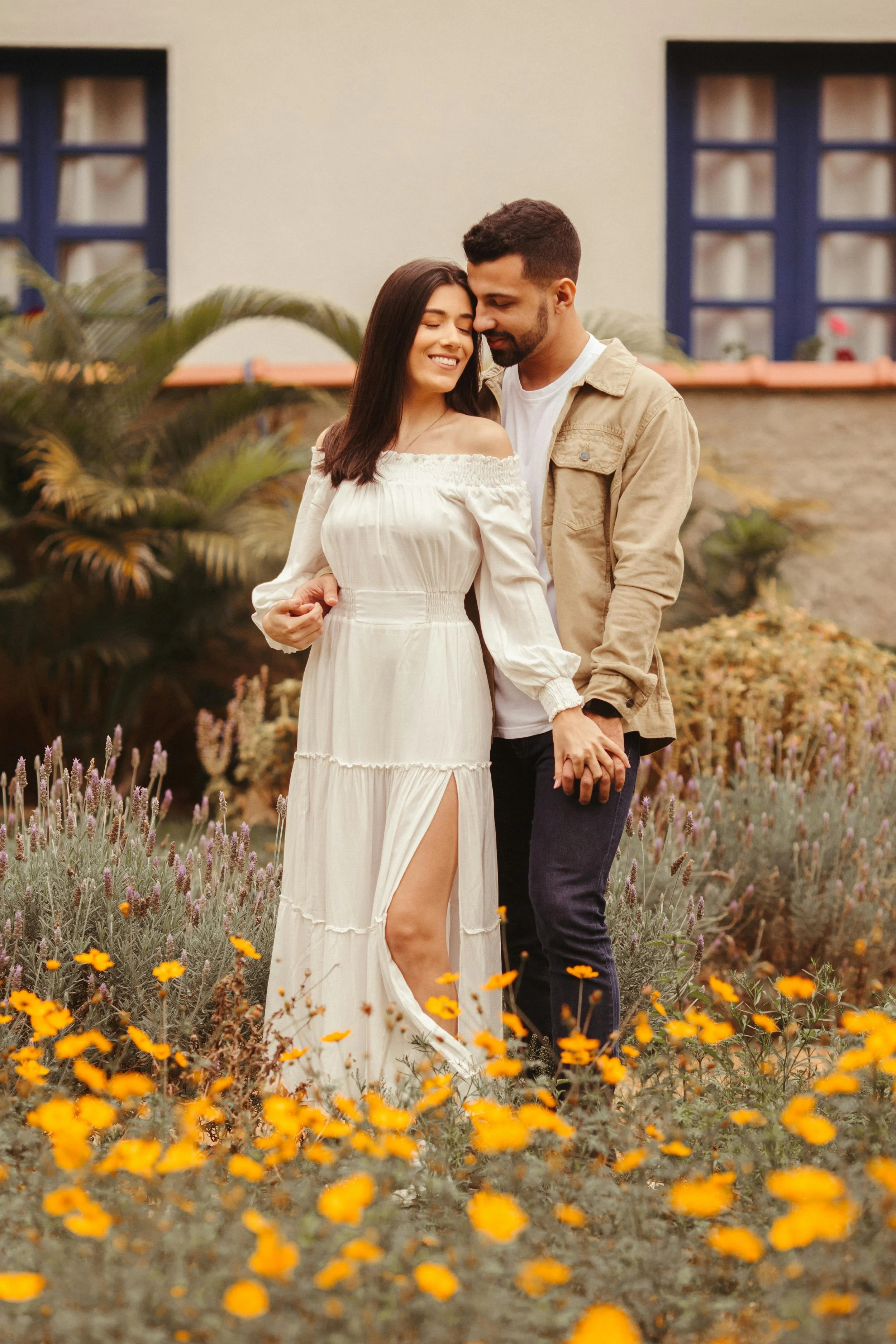 A young couple holding hands and smiling in a garden with yellow and purple flowers, with a building featuring blue window frames in the background.