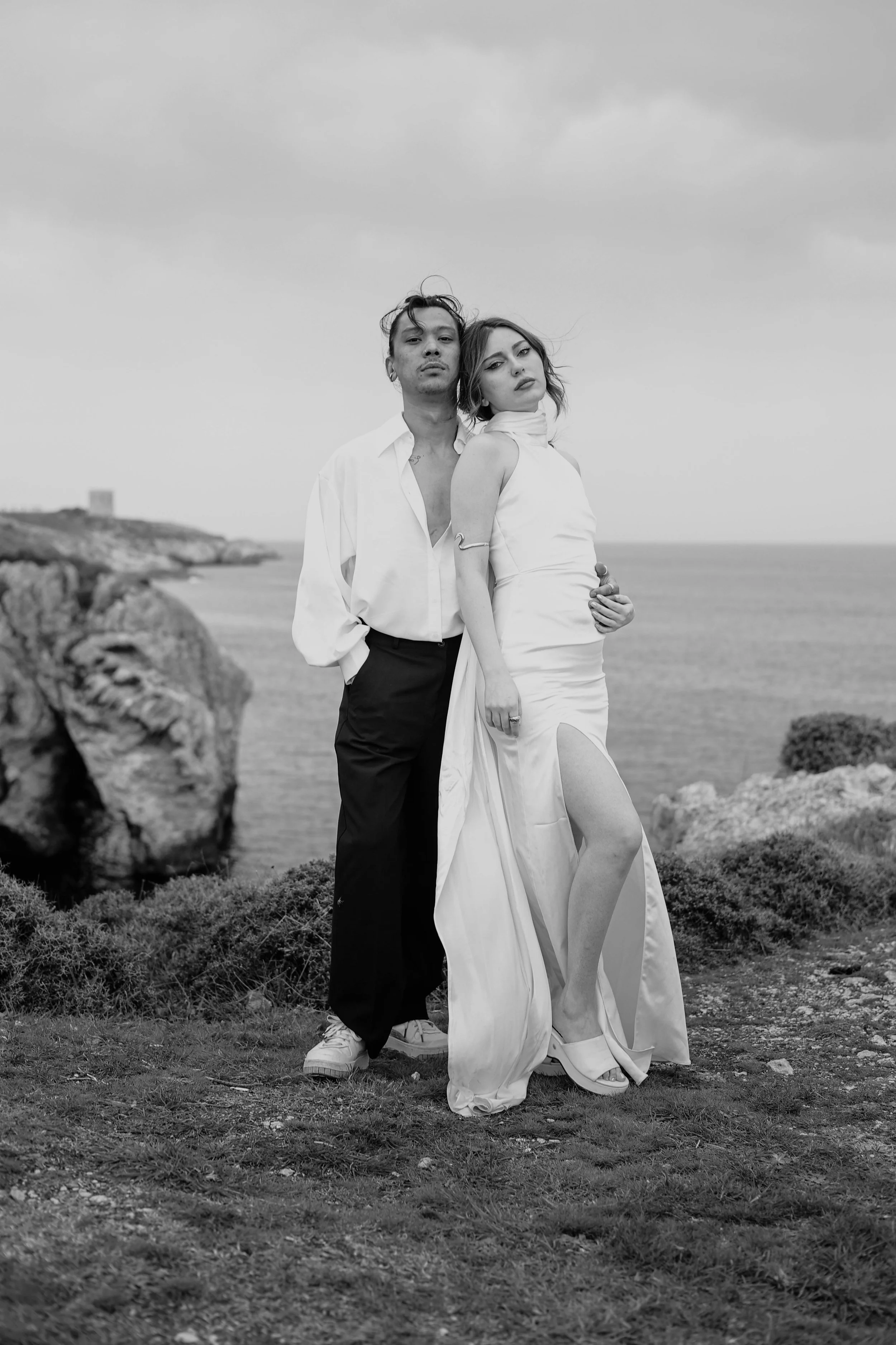 Black and white photo of a man and woman standing close together by the coastline. The woman is wearing a sleeveless evening gown with a high slit, and the man is dressed in a white shirt and dark pants. They are looking at the camera with serious expressions, with rocks and the ocean in the background.