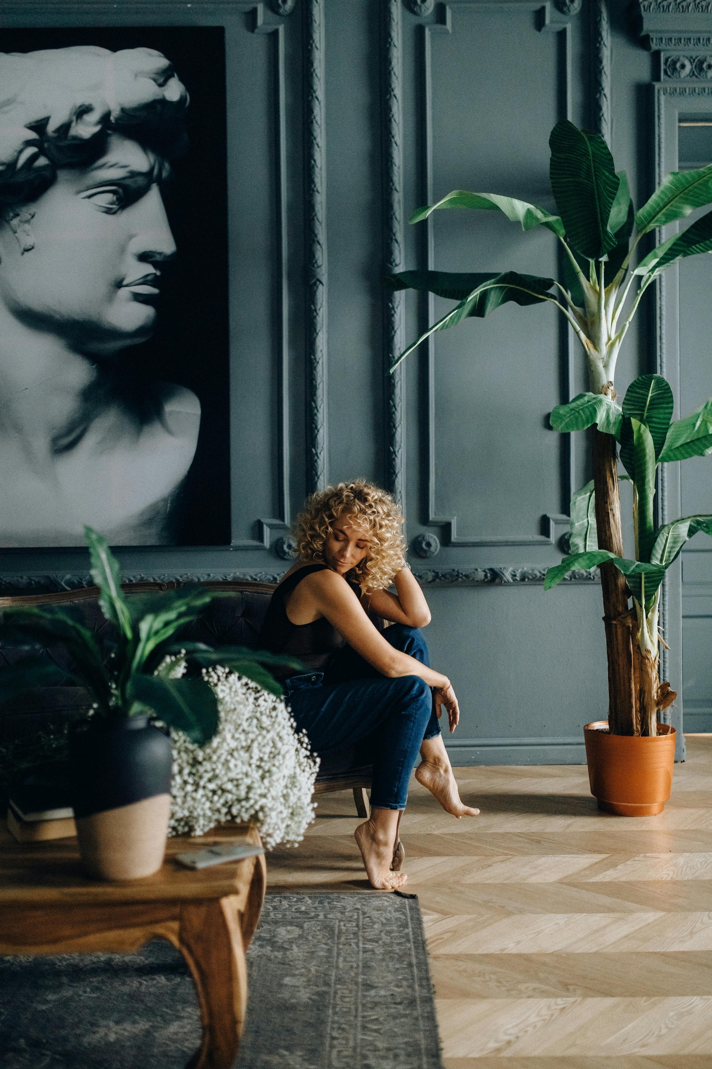 A woman with curly blonde hair sitting on a dark leather sofa in a room with dark gray walls, large plant, and black and white classical bust painting.