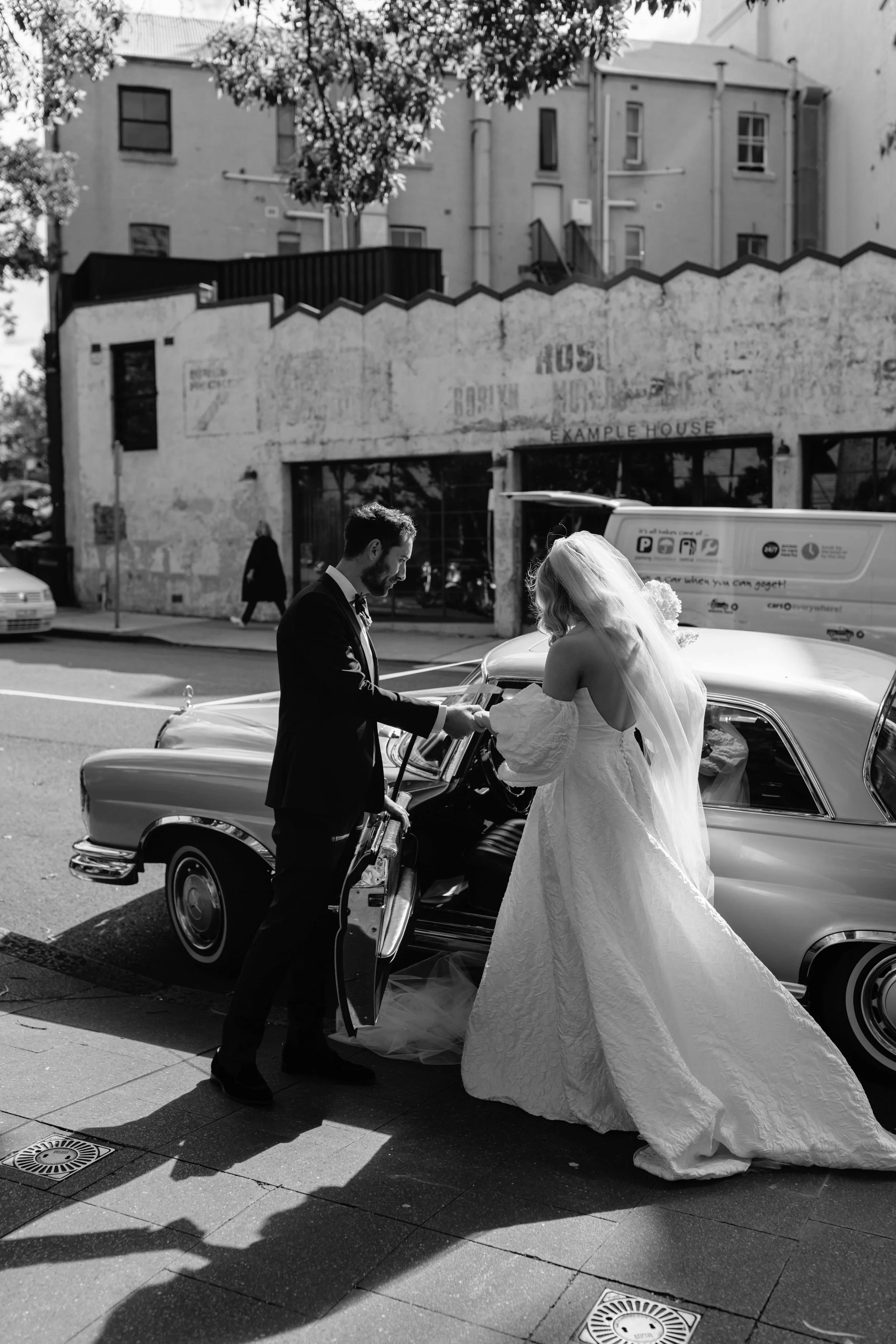 A bride and groom exchanging rings beside a vintage car on a city sidewalk.