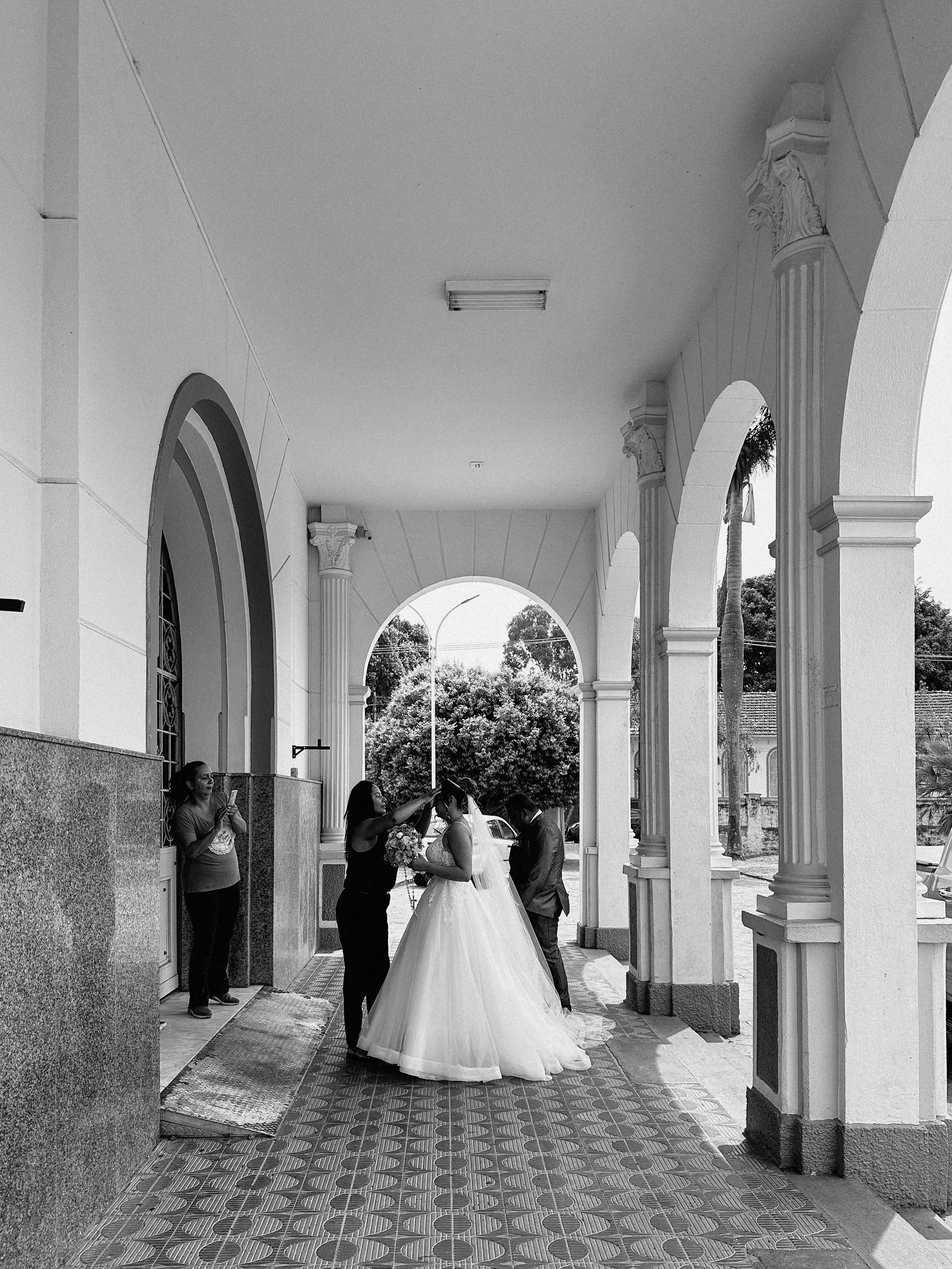 A bride in a white wedding gown is getting ready, assisted by a woman, with a man standing nearby. They are under a covered outdoor porch with archways and columns, with two women on the side, one taking a photo with her phone. There are trees and parked cars in the background.