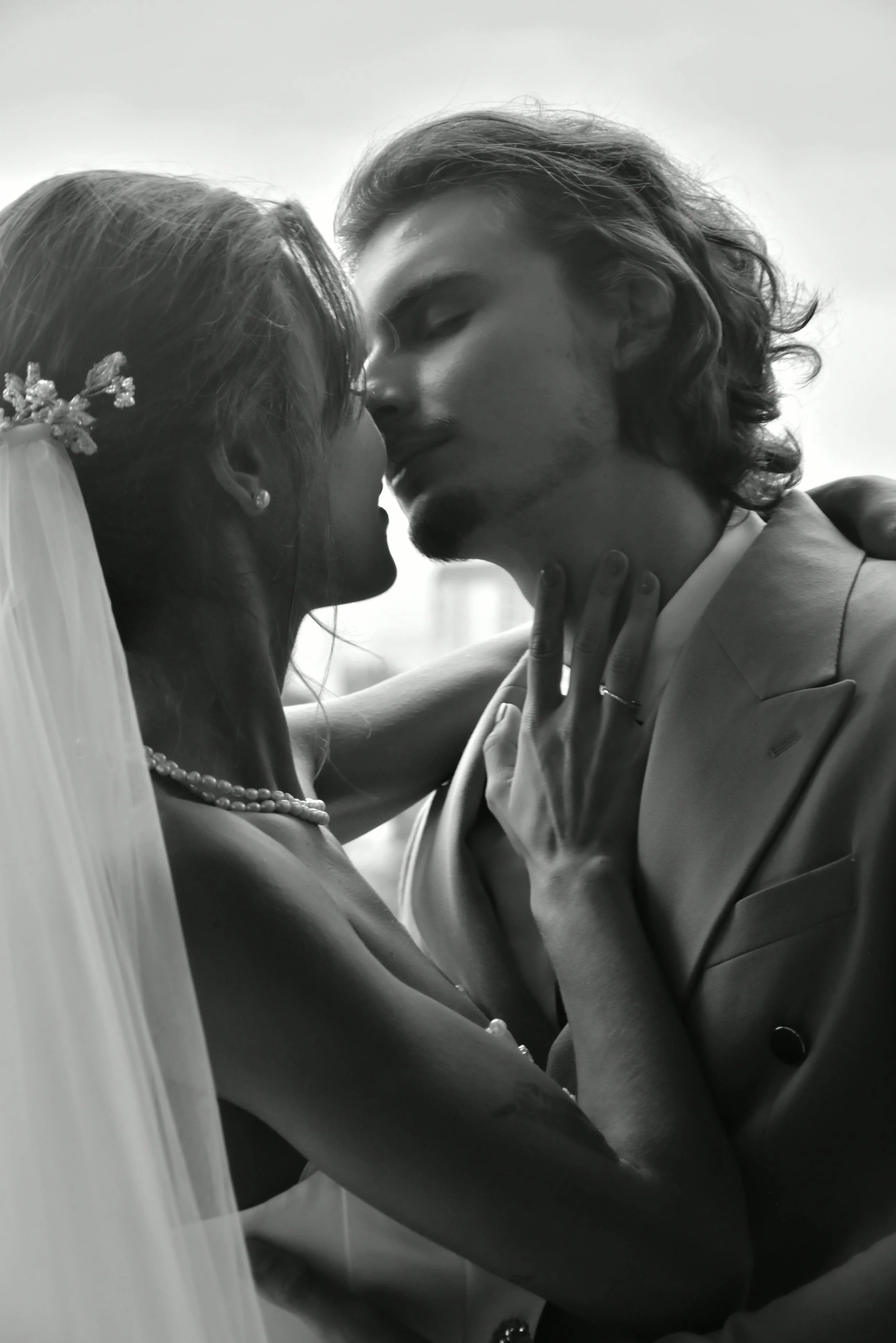 A black and white photo of a bride and groom kissing closely, with the bride wearing a veil, pearl necklace, and earrings, and the groom wearing a suit.