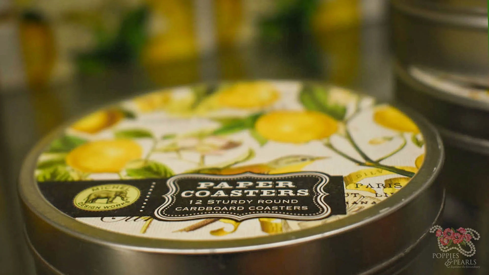 Round tin of paper coasters with a label that reads 'Paper Coasters, 12 Sturdy Round Cardboard Coasters' on top of yellow and green lemon-themed paper coaster.