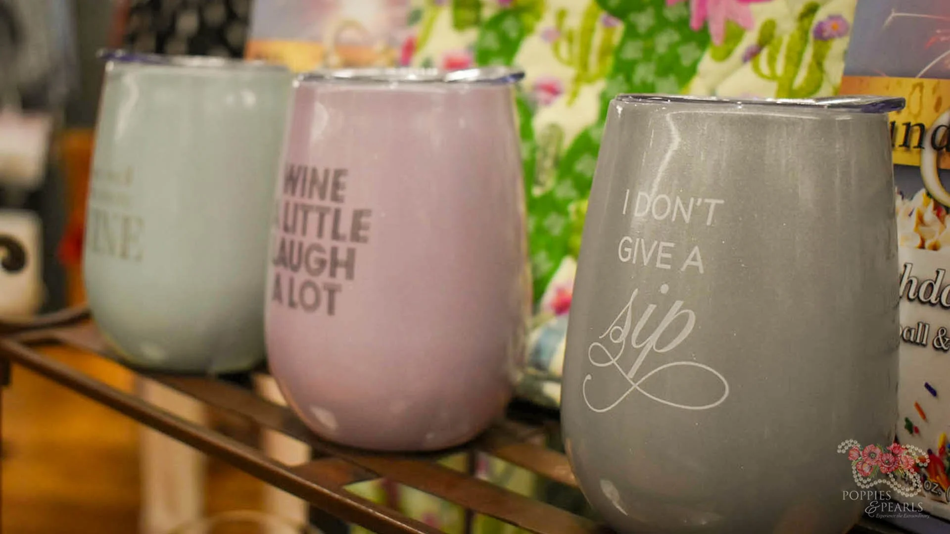 Three pastel-colored stemless wine glasses with humorous phrases on them, displayed on a wooden rack in a store. Each glass has a unique phrase: "I don't give a sip" on the gray glass, "Wine little, laugh a lot" on the pink glass, and the third glass