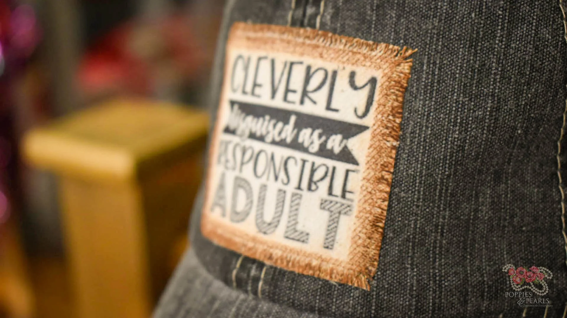 Close-up of a denim fabric with a sewn patch that says, "Cleverly disguised as an responsible adult." The background is blurred, showing a hint of purple and yellow.