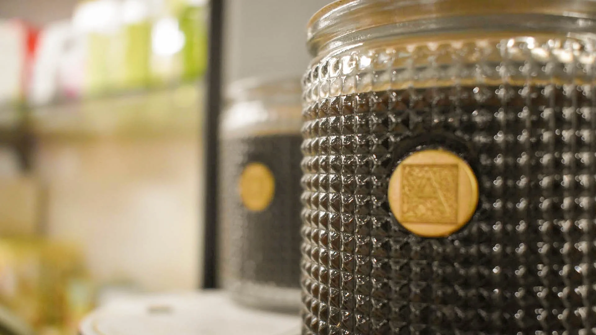 Close-up of a decorative glass jar with a gold emblem, containing dark coffee beans, in a kitchen or store setting.