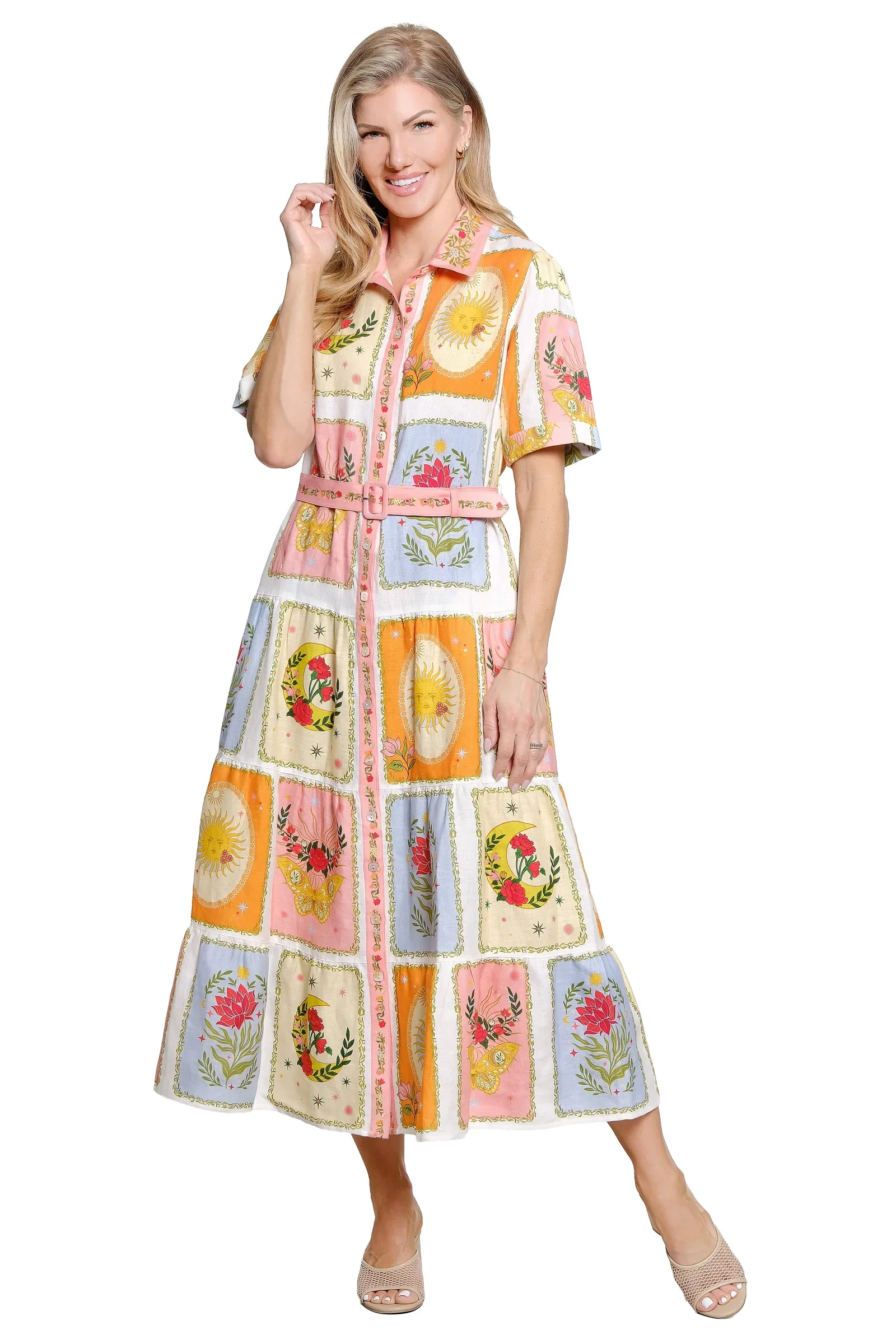 A woman wearing a colorful, patterned, short-sleeve maxi dress with a pink belt, standing and smiling with one hand touching her hair on a white background.