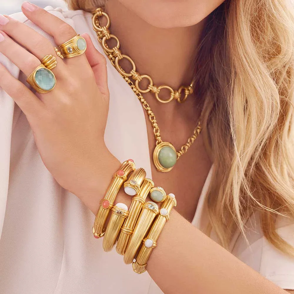 A woman wearing gold jewelry, including multiple rings, two necklaces with large circular green stones, and a wide gold bracelet with pink, white, and clear stones.