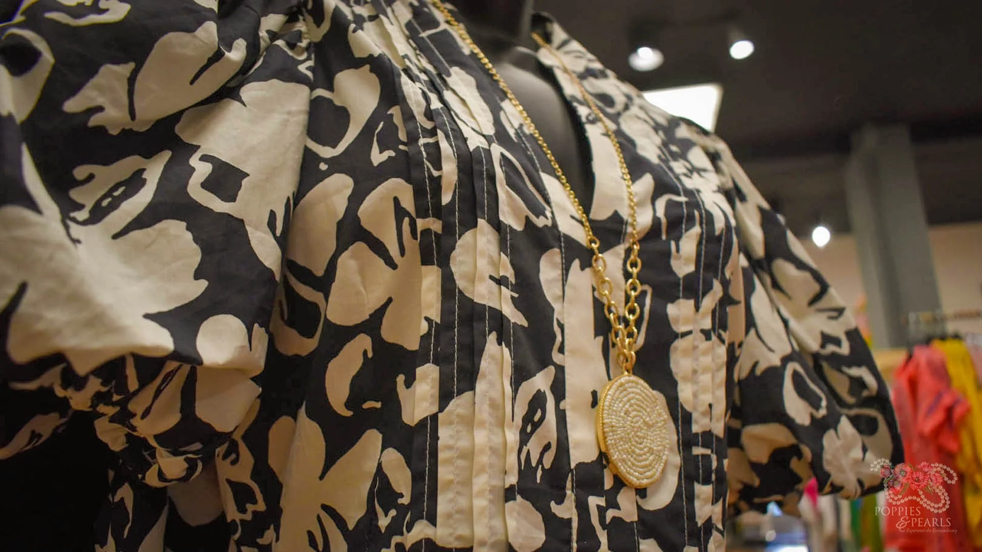 A mannequin dressed in a black and white patterned blouse, accessorized with a long gold chain necklace featuring a large oval pendant.