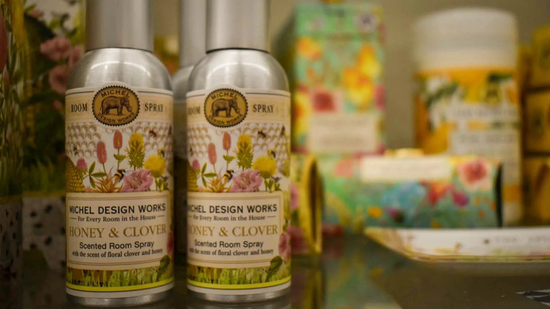 Two bottles of Michel Design Works honey and clover scented room spray on a store shelf, with colorful packaging in the background.
