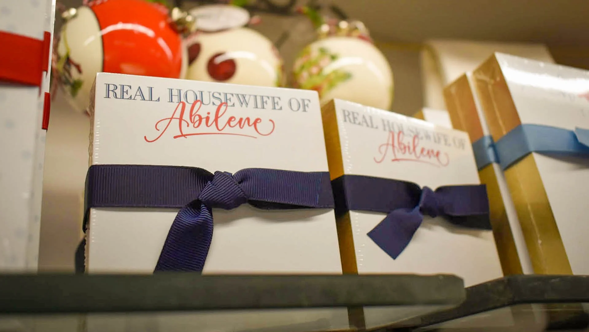 Gift boxes with text 'Real Housewife of Abilene' and colored ribbons, set on a shelf with decorative ceramic ornaments in the background.