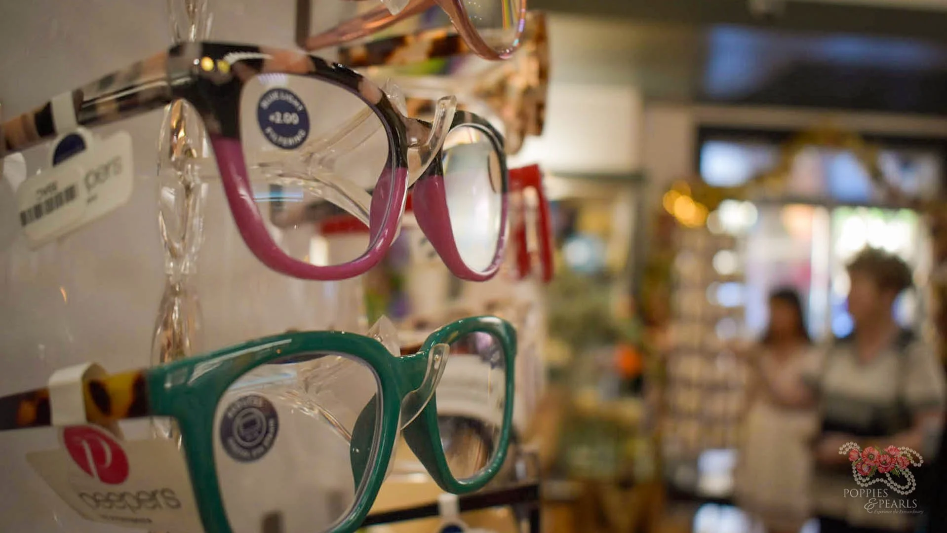Colorful eyeglasses on display shelf in a store, with two women shopping in the blurry background.