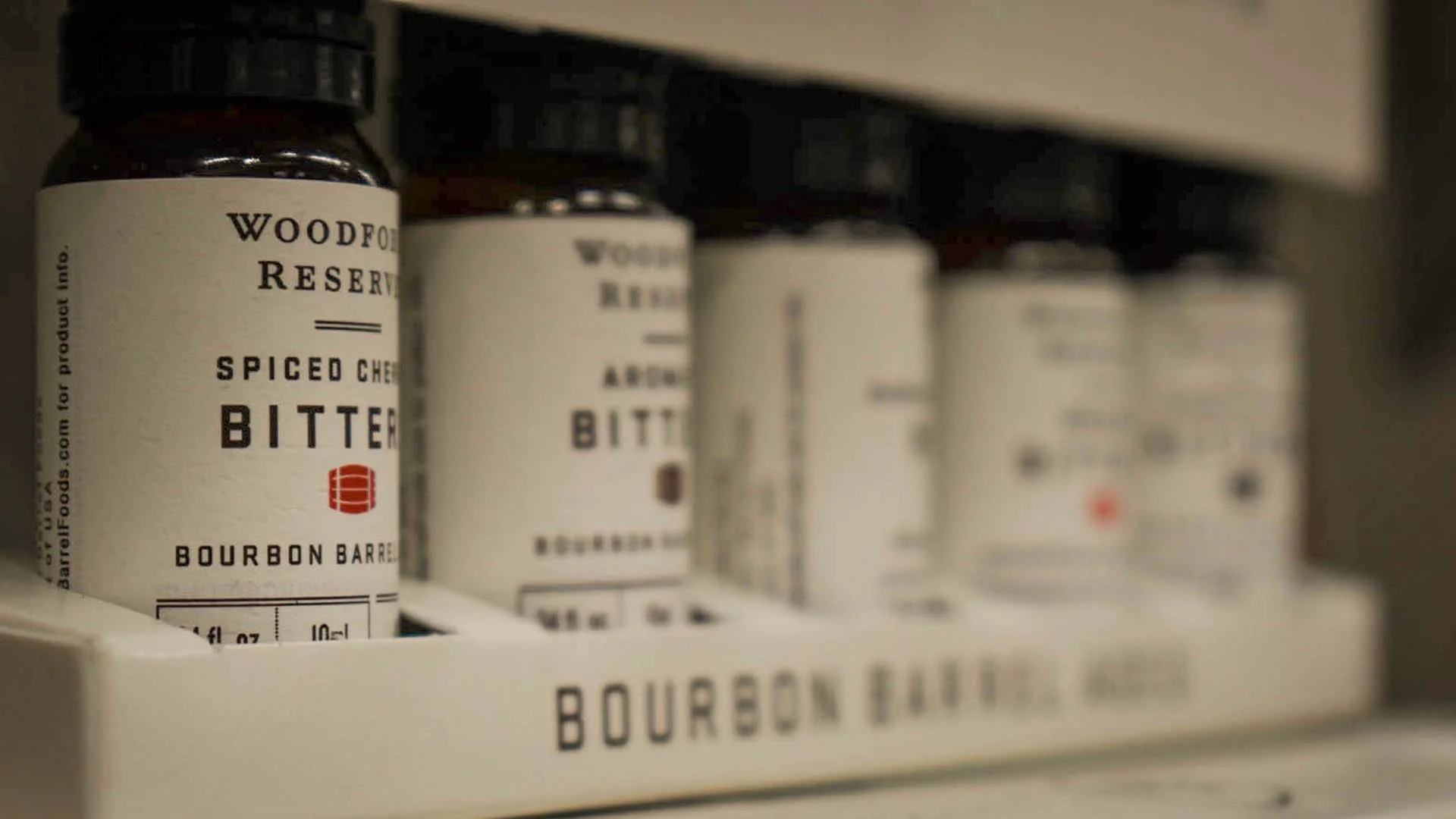 Close-up of a shelf with bottles of Woodford Reserve bourbon bitters, with other bottles blurred in the background.