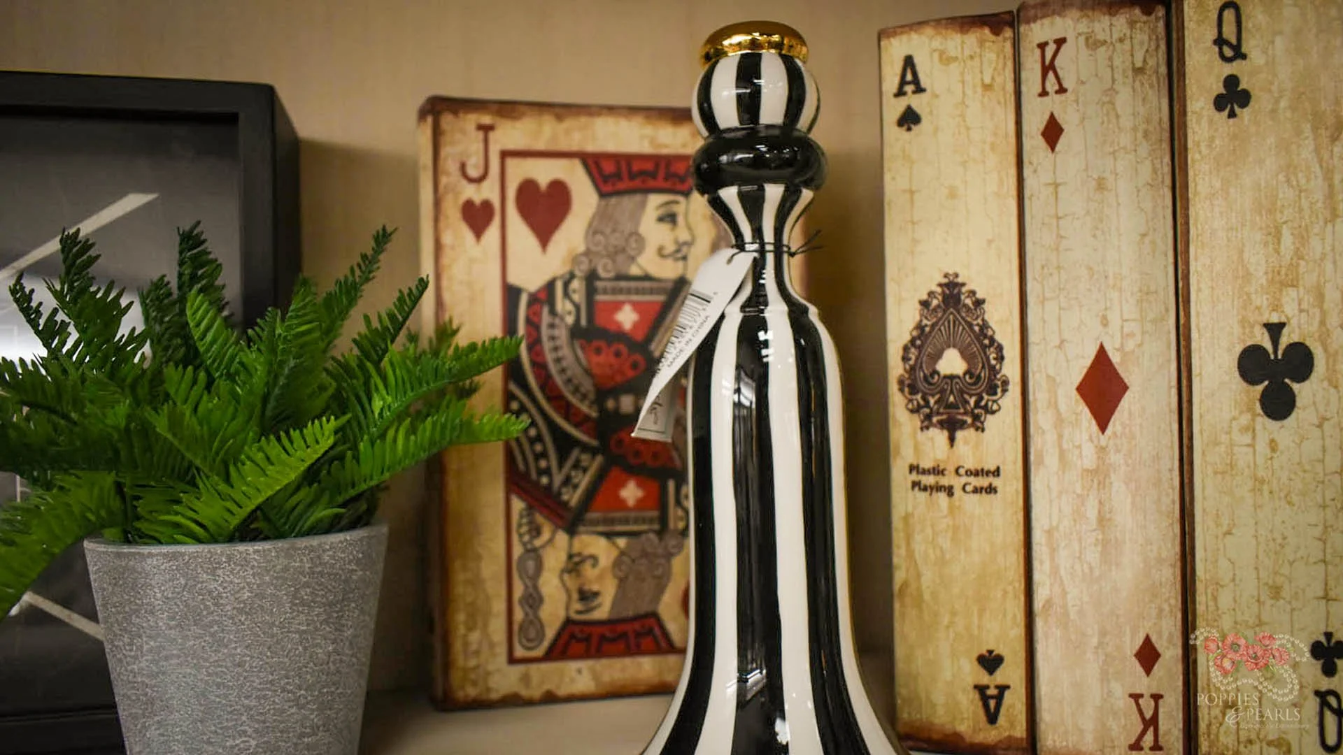 Decorative black and white striped glass bottle in front of vintage-style playing card boxes and a potted green fern plant.