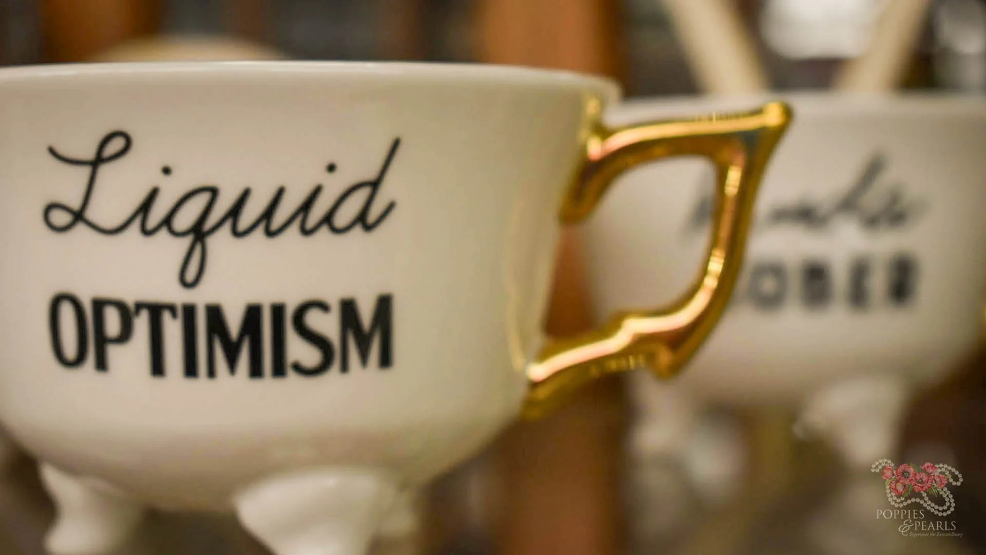 Close-up of two white coffee cups with black text, one says 'Liquid OPTIMISM' with a gold handle in the shape of a letter D. The other cup is partially visible and out of focus.