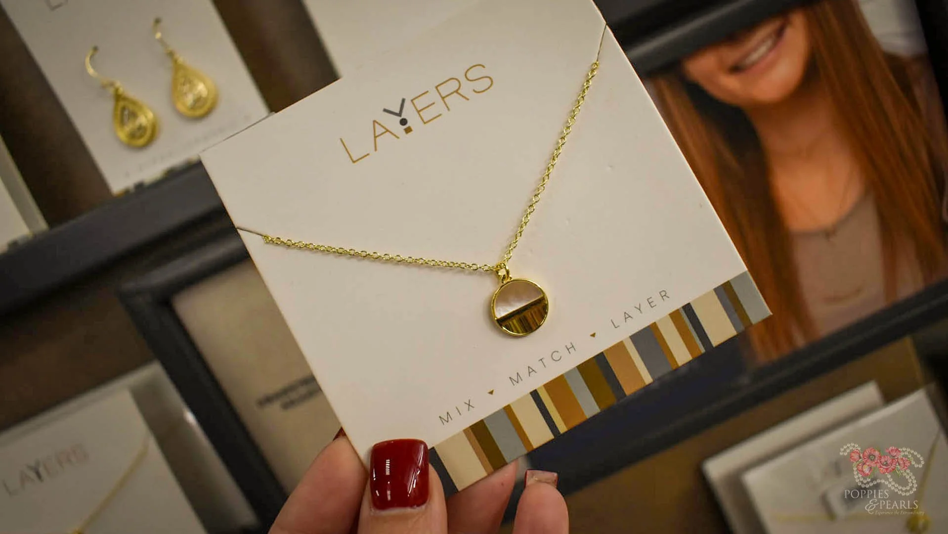 A gold layered necklace with a circular pendant displayed on a white card that says 'LA:ERS' in a jewelry store. The card also has the words 'MIX MATCH LAYER' at the bottom. The person holding the card has red painted nails.
