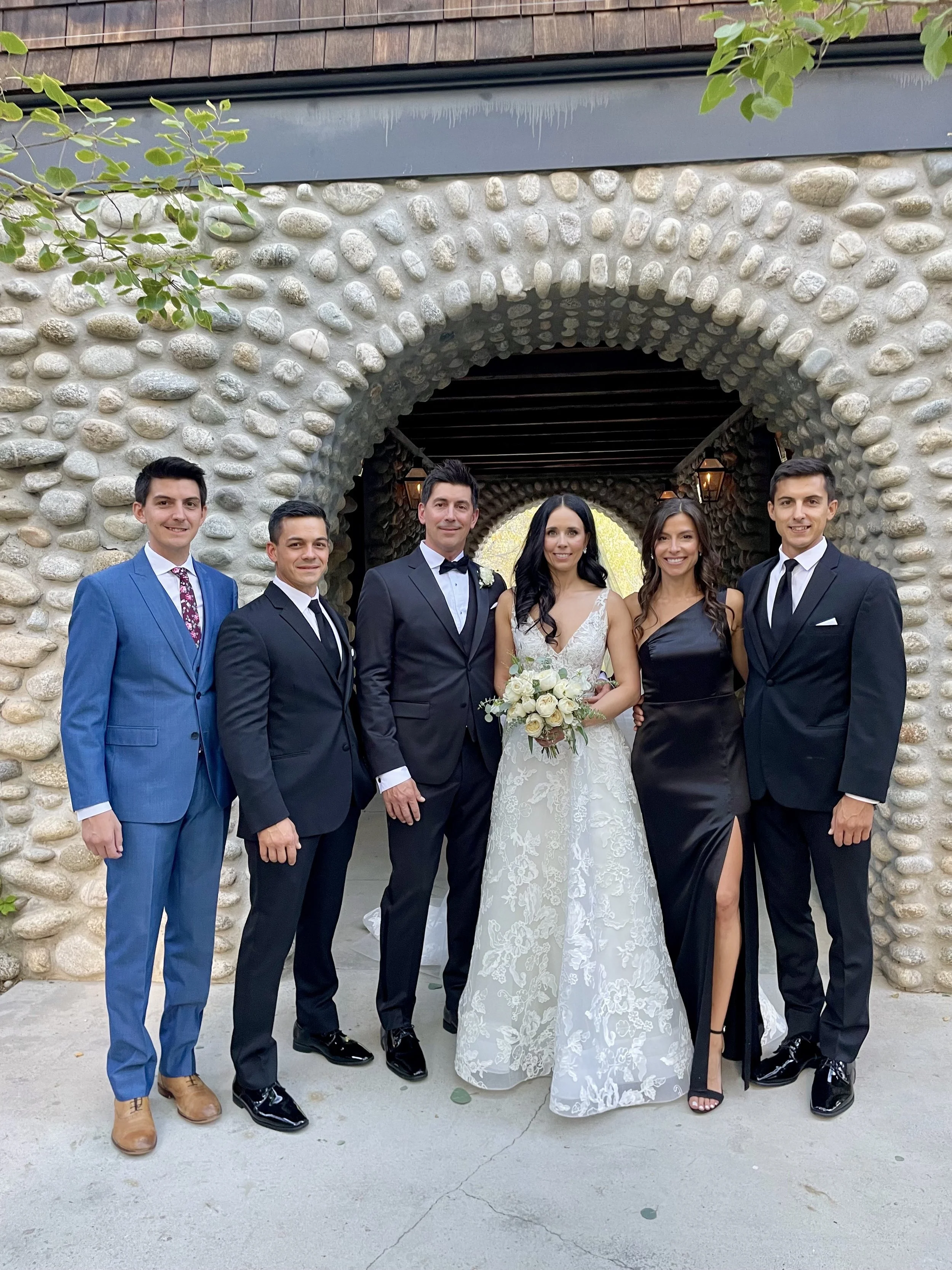 Group of six people in formal wedding attire standing under a stone wedding ceremony archway outside, with the bride holding a bouquet of white flowers, and groom next to the bride, all smiling at the camera.