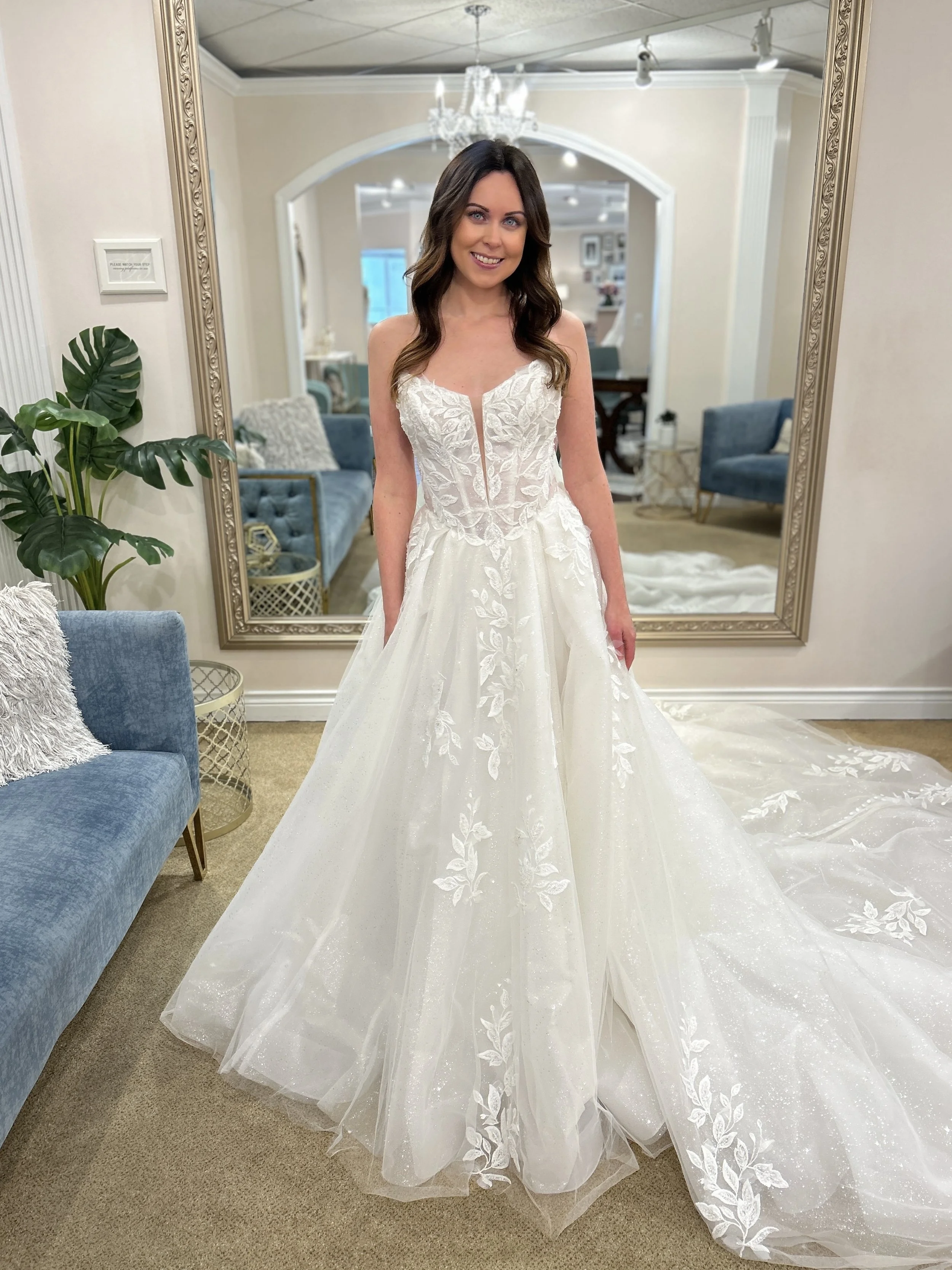 Woman in a white, ivory strapless corset ballgown wedding dress with floral embroidery and sparkle standing in a fitting room with a large mirror, chandelier, and blue furniture.