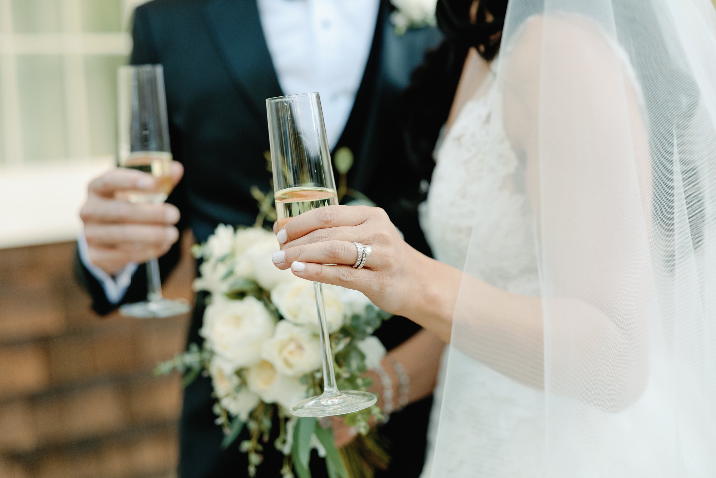 Close-up of bride and groom holding champagne flutes during wedding toast, with bride wearing a veil and wedding dress, groom in suit, and bride holding a bouquet of white flowers.