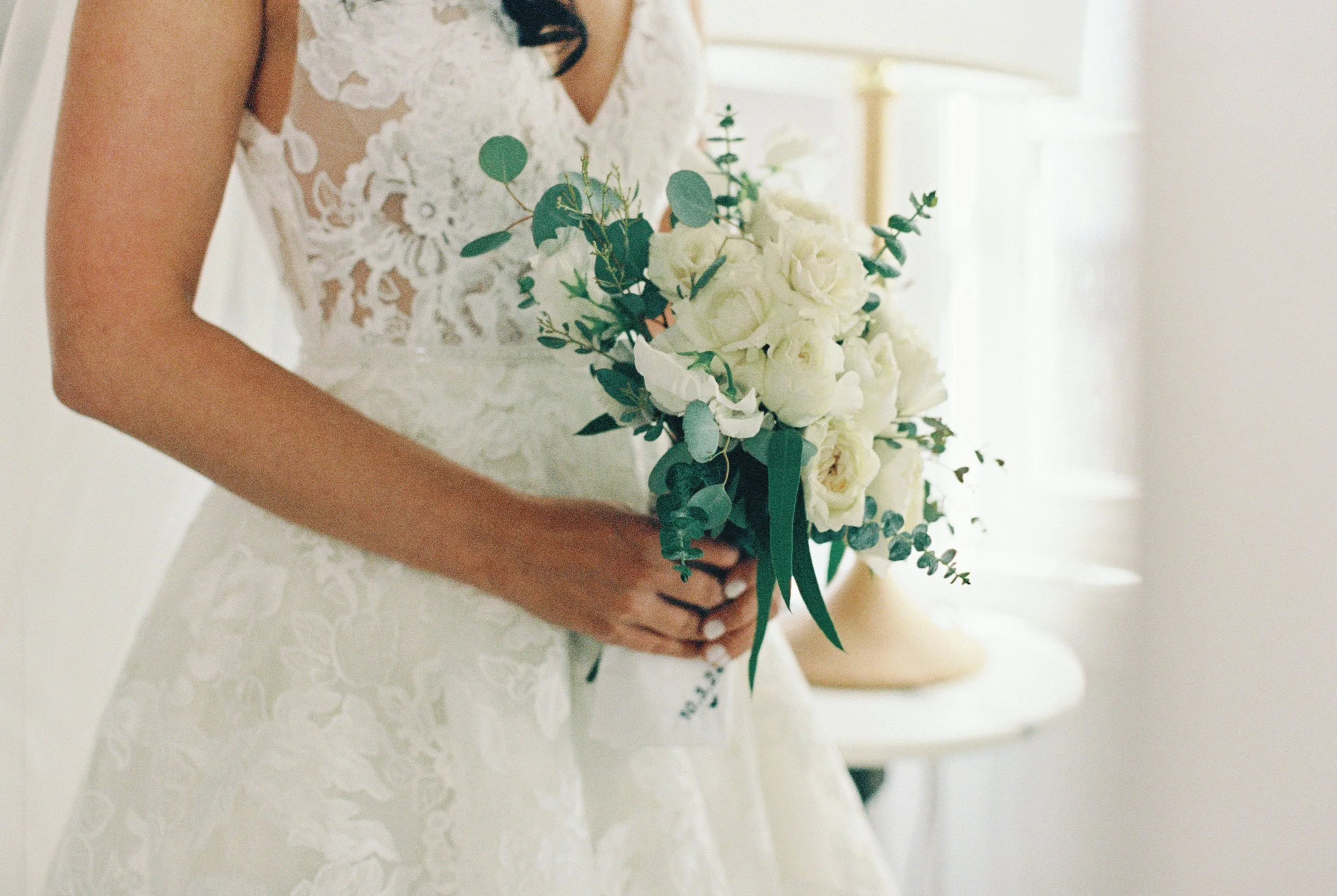 A woman in a lace wedding dress holding a bouquet of white roses and greenery.