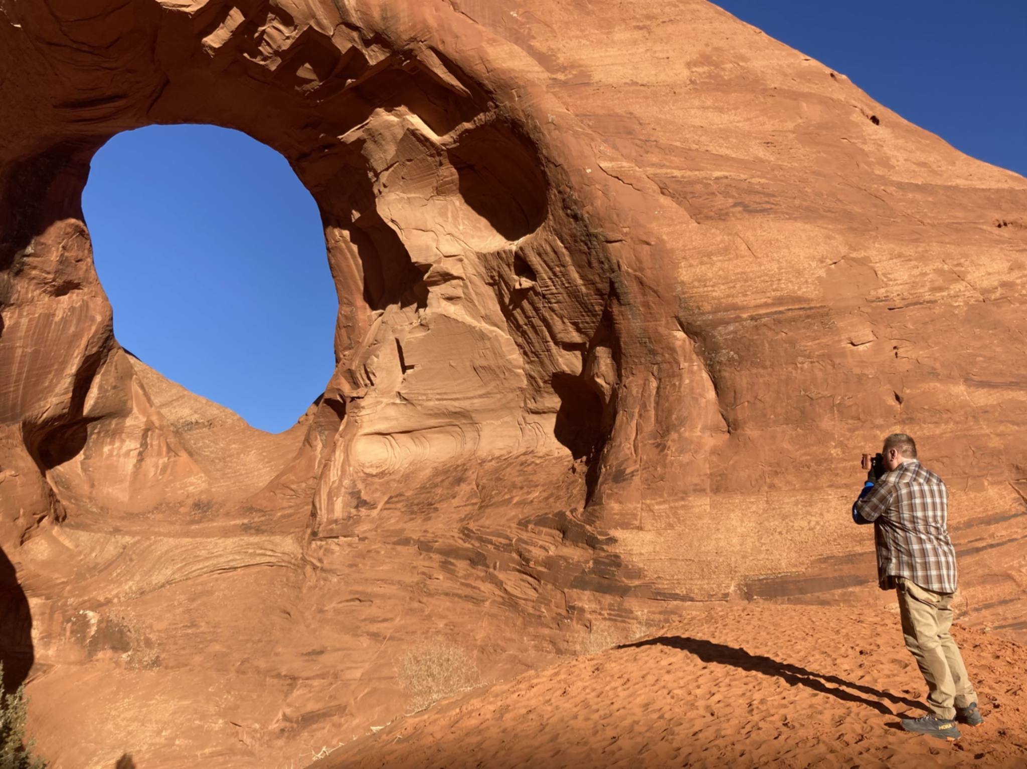 A man in a plaid shirt and khaki pants stands on sandy desert ground, taking a photo of a large, naturally eroded red sandstone rock formation with a hole in the middle, against a blue sky.