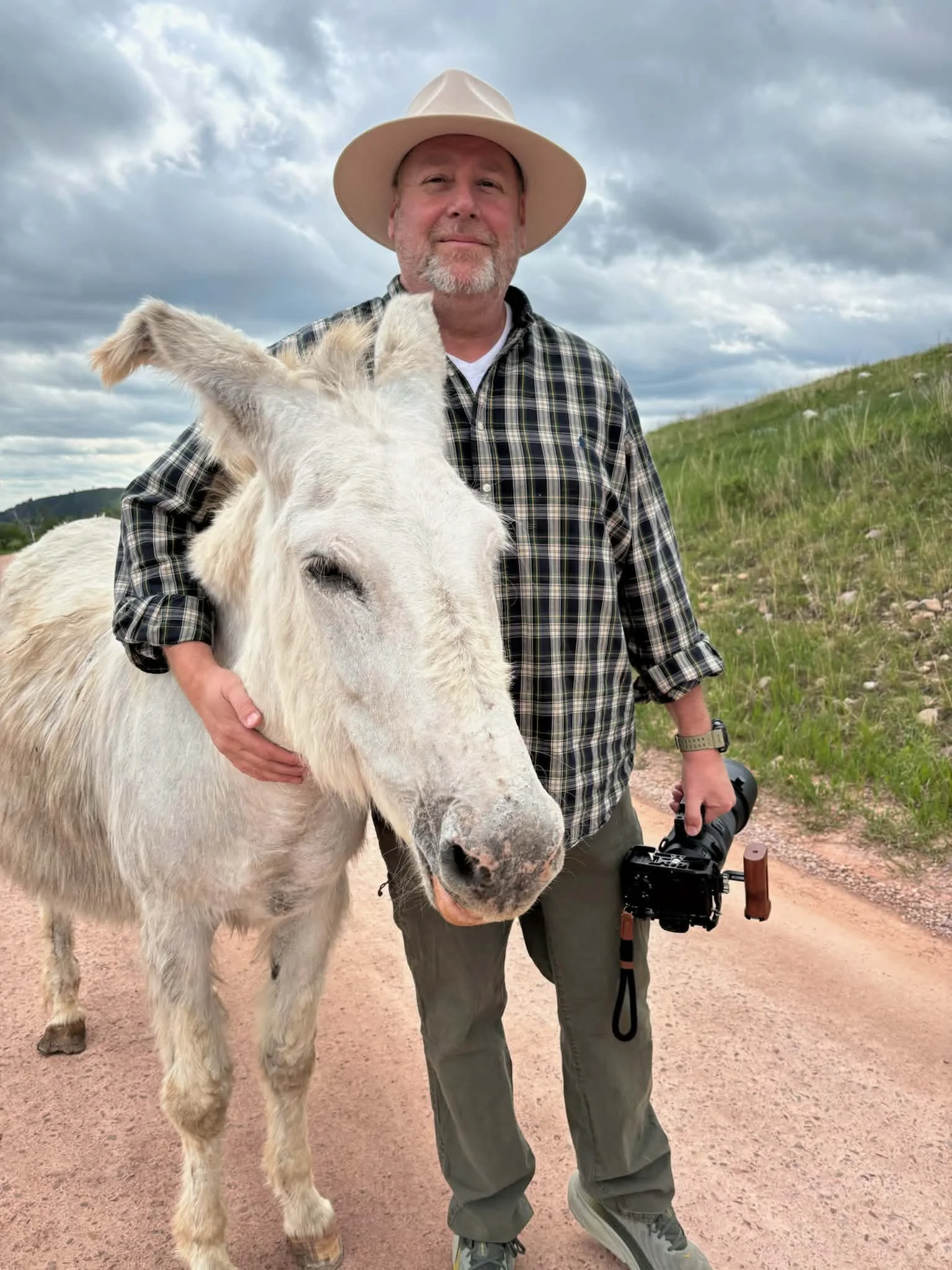 A man in a plaid shirt and a wide-brimmed hat standing outdoors on a dirt path, holding a camera in his right hand, next to a white donkey with a friendly expression.