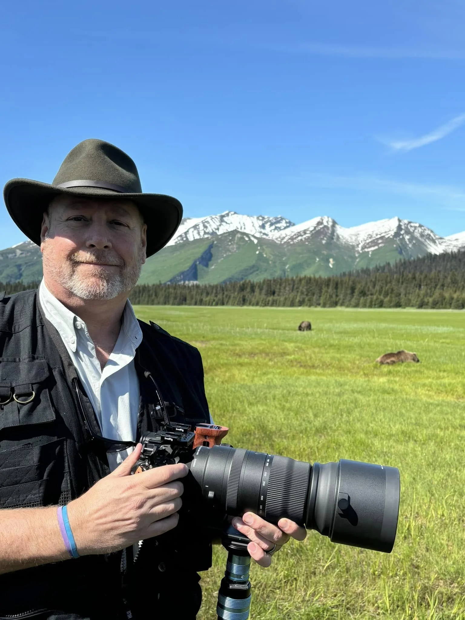 A man wearing a brown wide-brimmed hat and a white shirt holding a professional camera with a large telephoto lens, standing in a green field with mountains and snow in the background.